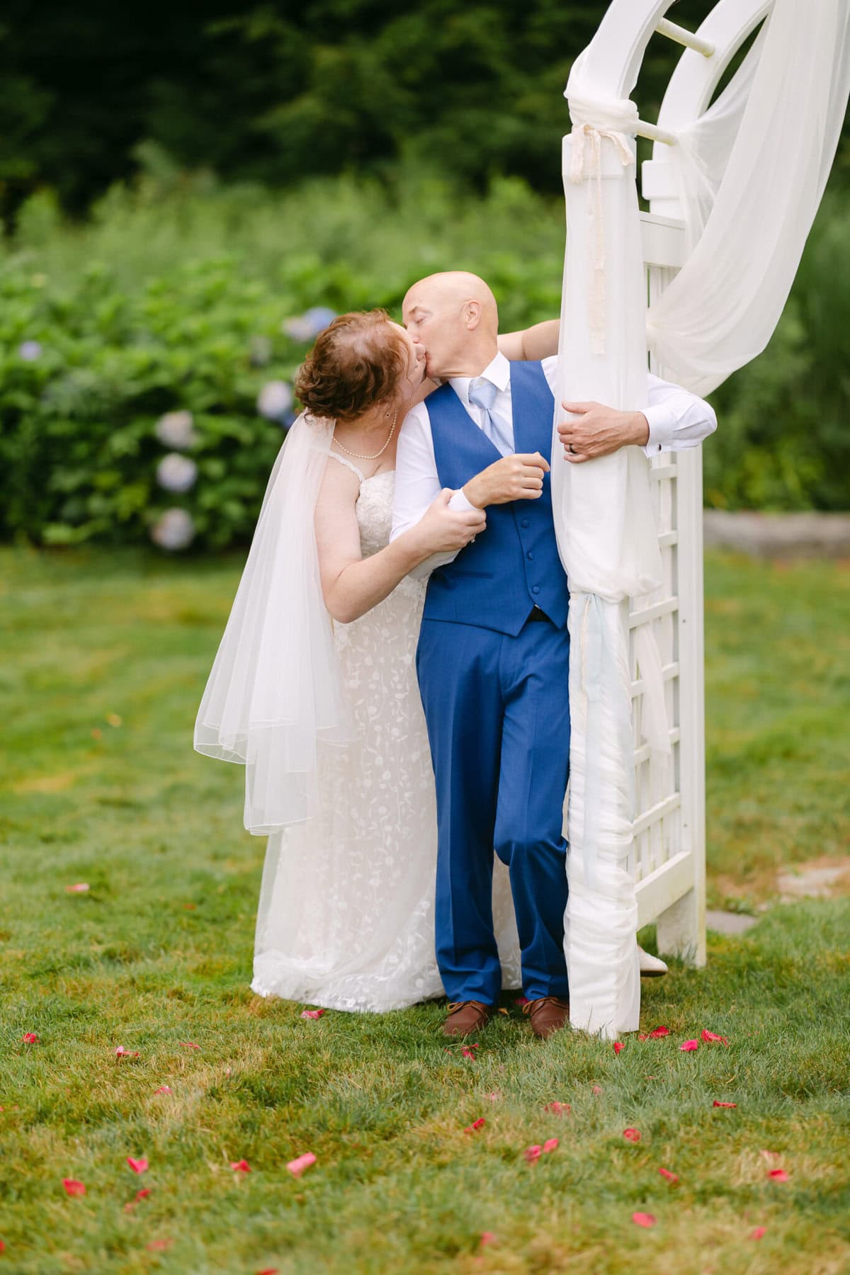 Wedding At the Holliston Historical Society 60 A bride and groom share a kiss under a white arch at the Holliston Historical Society, with rose petals scattered across the wedding lawn.