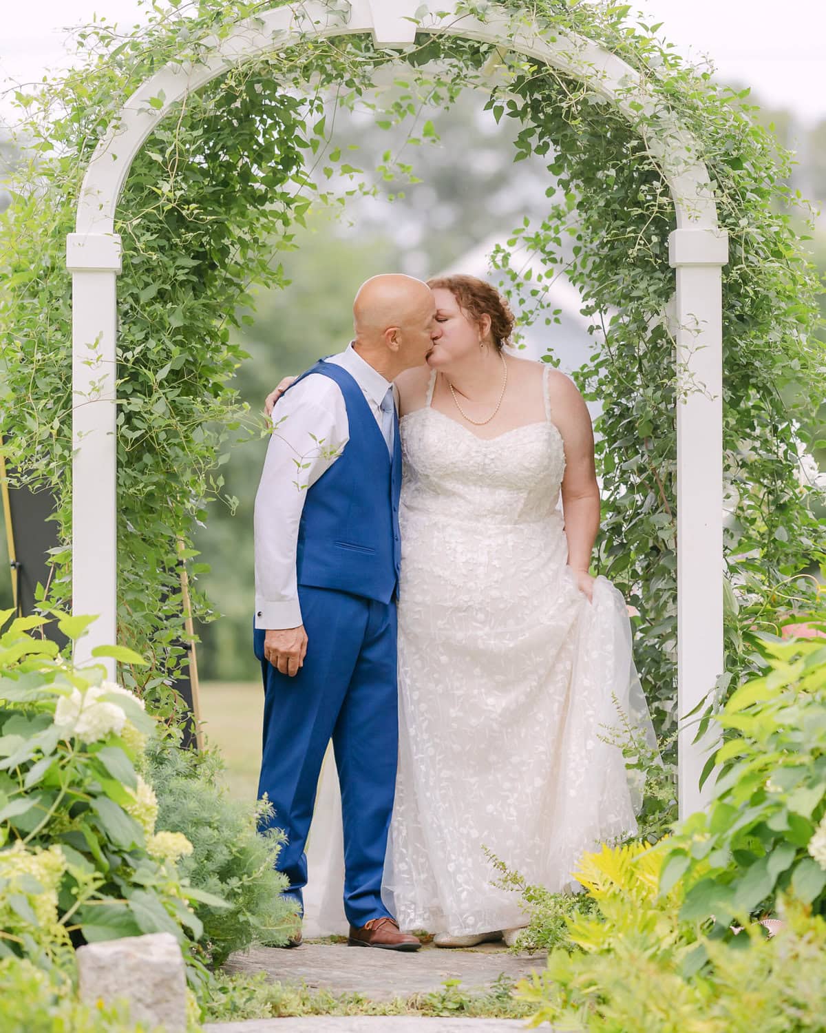 Wedding At the Holliston Historical Society 65 A bride and groom kiss under a white archway with green vines at a Holliston Historical Society wedding in an outdoor garden setting.