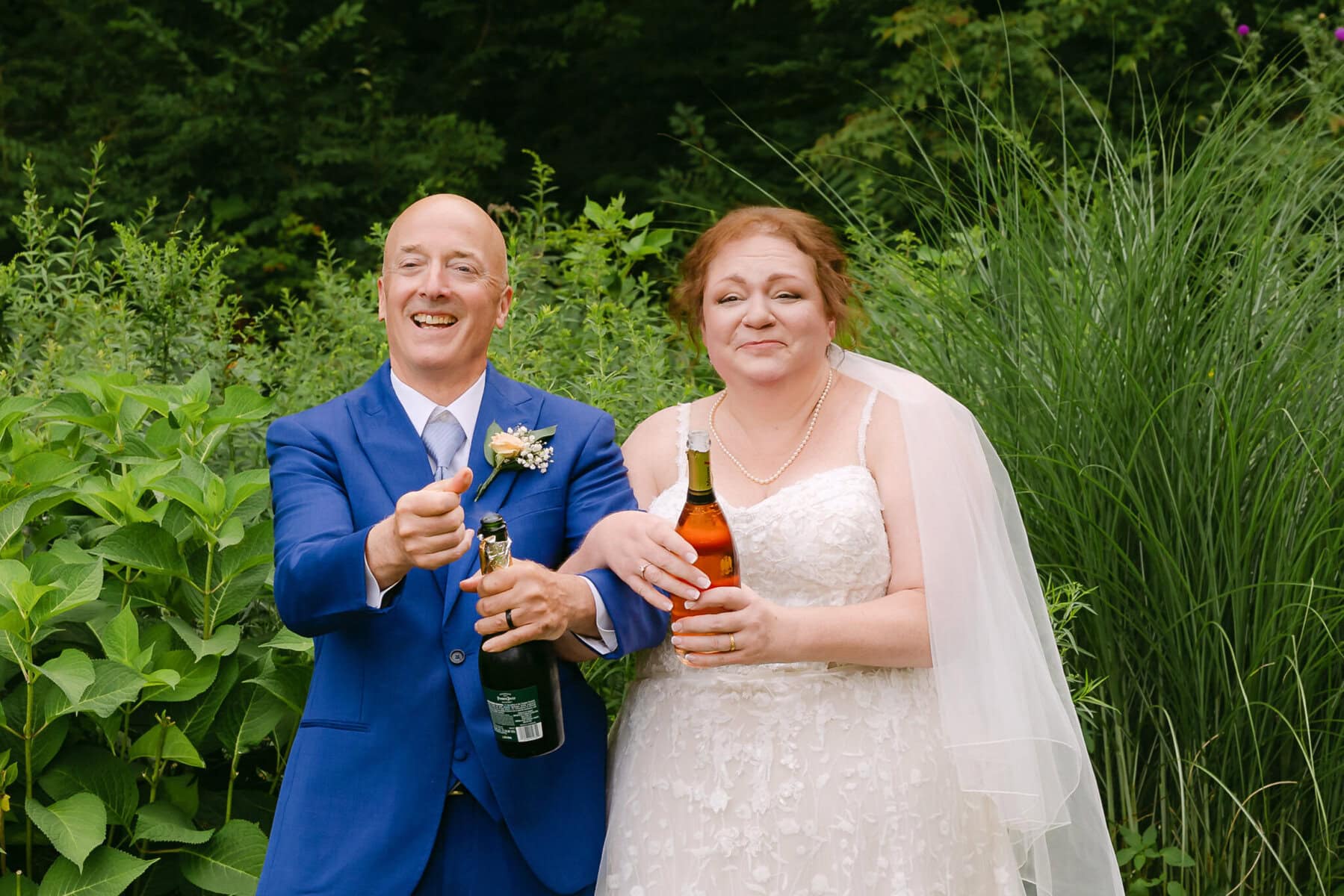 Wedding At the Holliston Historical Society 63 A bride and groom stand outdoors at the Holliston Historical Society, smiling and opening champagne amid lush wedding greenery.