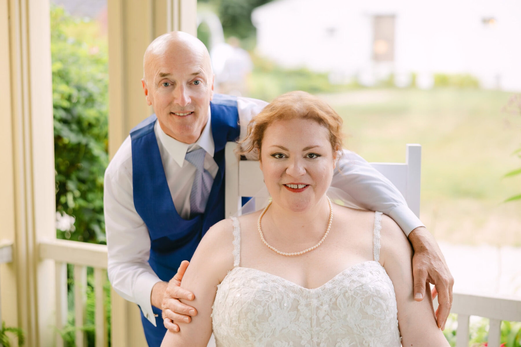 Wedding At the Holliston Historical Society 64 A man in a blue vest stands behind a bride in white lace at their wedding, both smiling on a porch at the Holliston Historical Society.