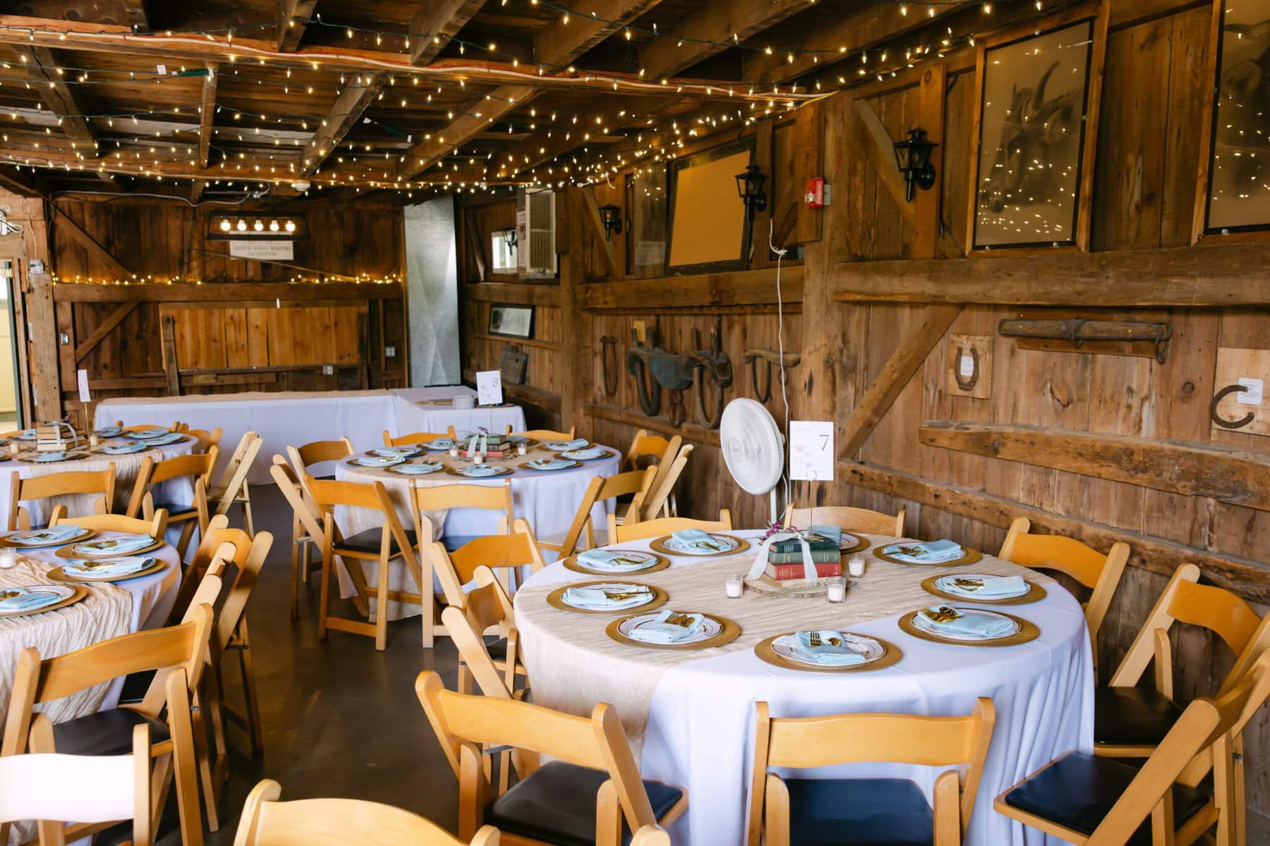 Wedding At the Holliston Historical Society 37 Wooden barn interior at Holliston Historical Society, with round tables set for a wedding, string lights, and rustic decor on the walls.
