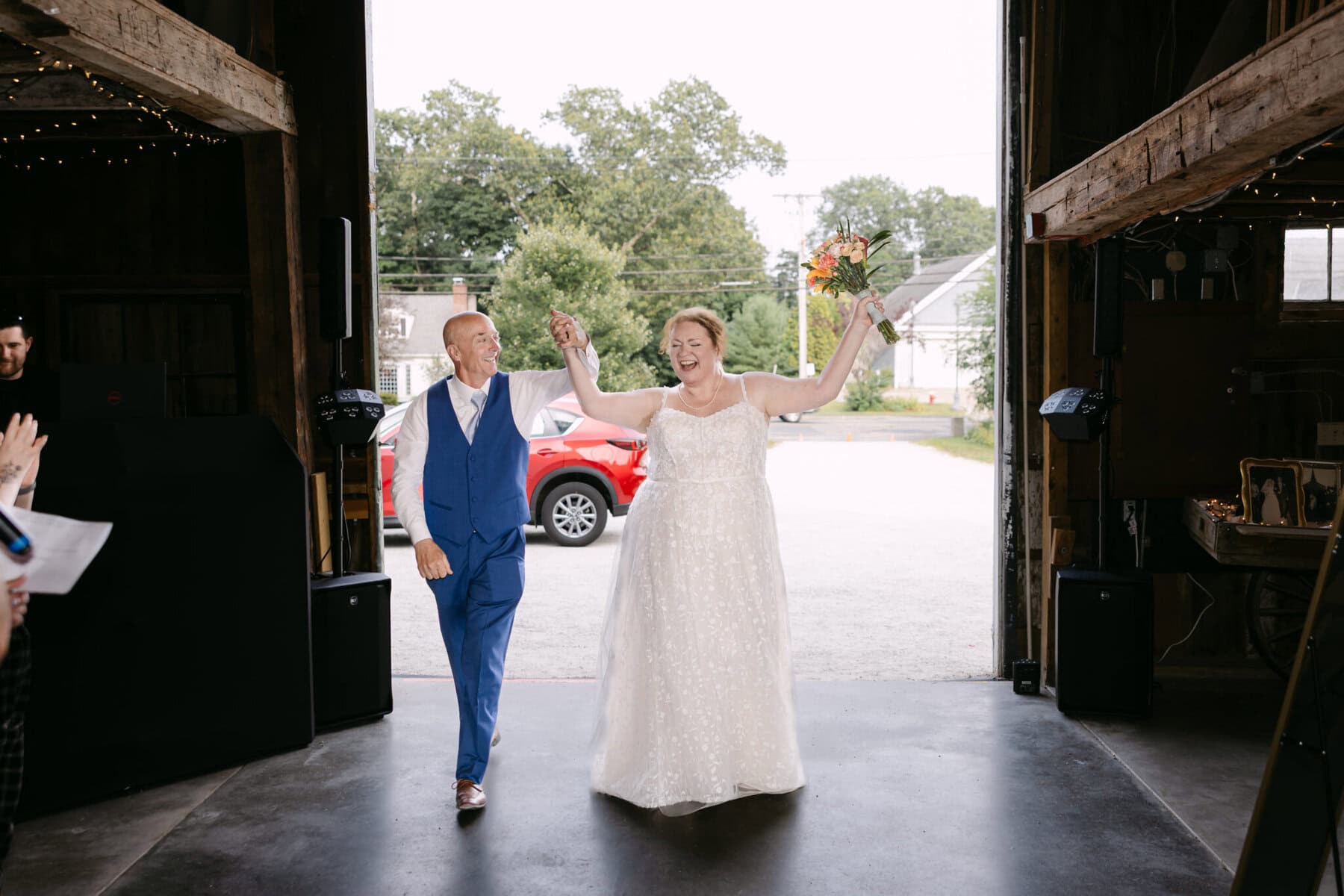 Wedding At the Holliston Historical Society 41 A bride in white and a man in blue enter the Holliston Historical Society barn, smiling with raised arms at their joyful wedding entrance.