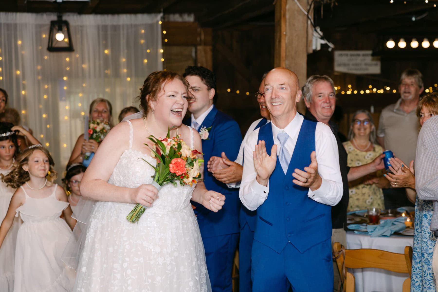 Wedding At the Holliston Historical Society 42 A bride holding flowers and a man in a blue vest clap and smile at their wedding reception at Holliston Historical Society.