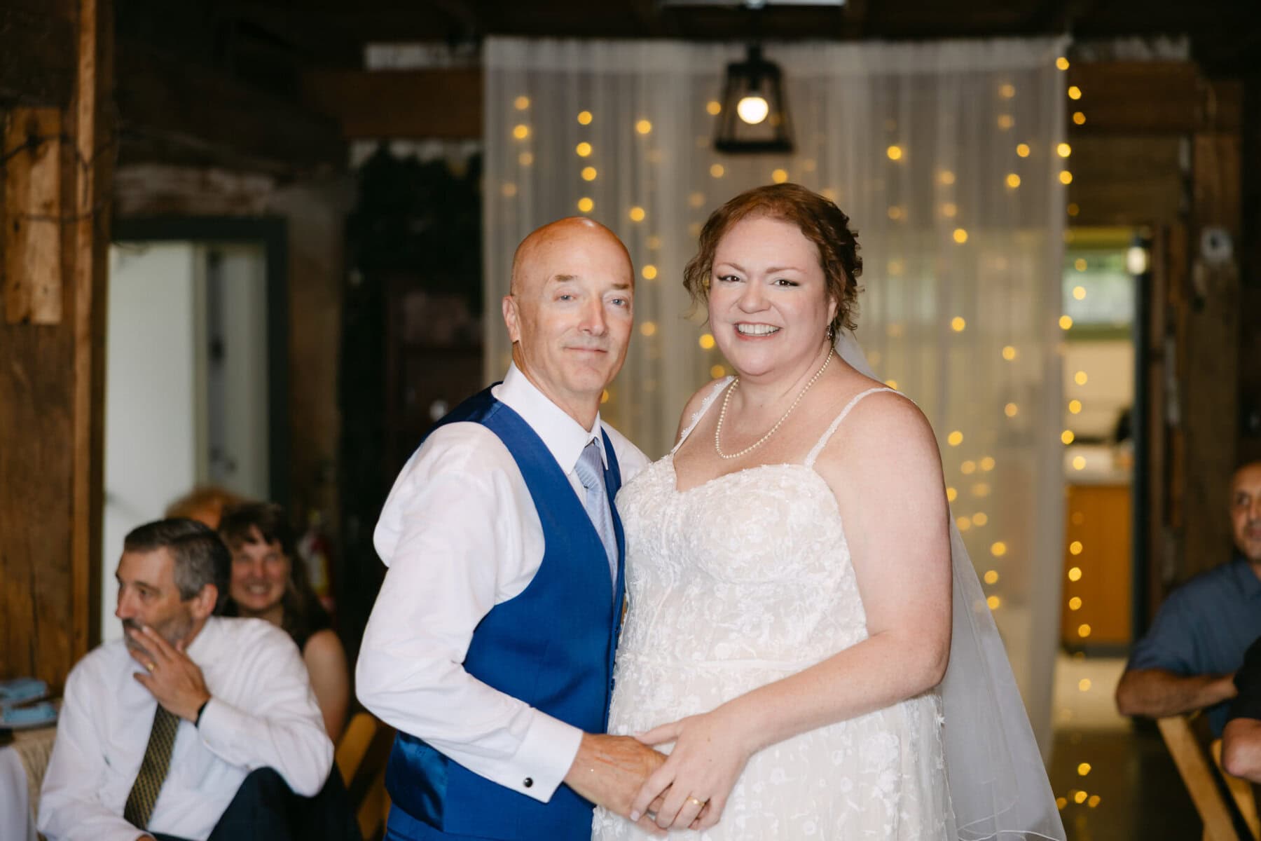 Wedding At the Holliston Historical Society 43 A bride and groom pose indoors at the Holliston Historical Society wedding, with glowing string lights in the background.