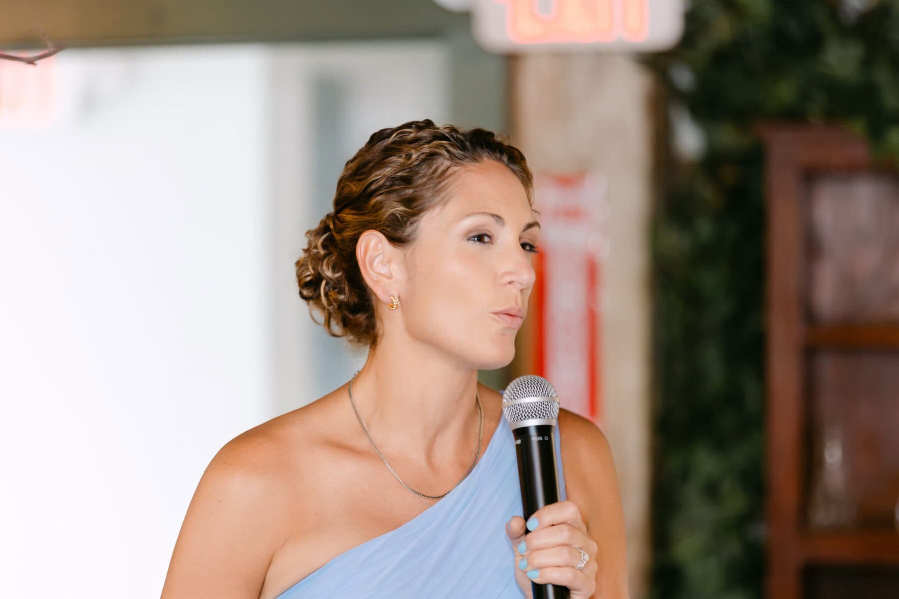 Wedding At the Holliston Historical Society 45 A woman in a light blue one-shoulder dress speaks into a microphone at a wedding inside the Holliston Historical Society.