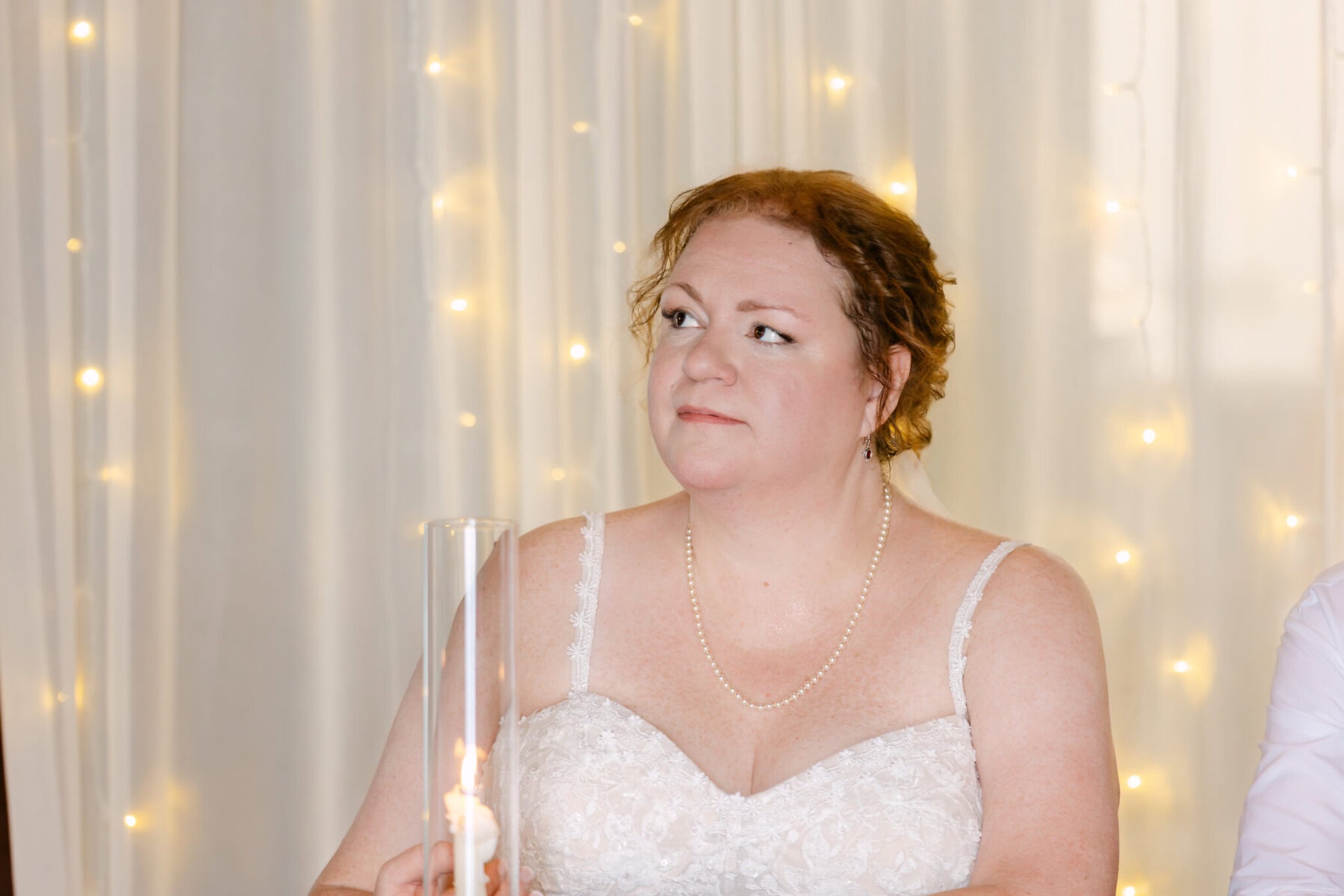 Wedding At the Holliston Historical Society 47 A woman in a white lace dress and pearl necklace sits before sheer curtains at a Holliston Historical Society wedding, holding a glass candle holder.