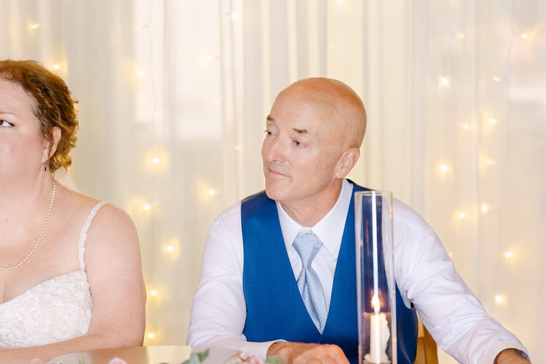 Wedding At the Holliston Historical Society 46 A man in a blue vest and tie sits beside a woman in a white wedding dress at the Holliston Historical Society, with string lights in the background.