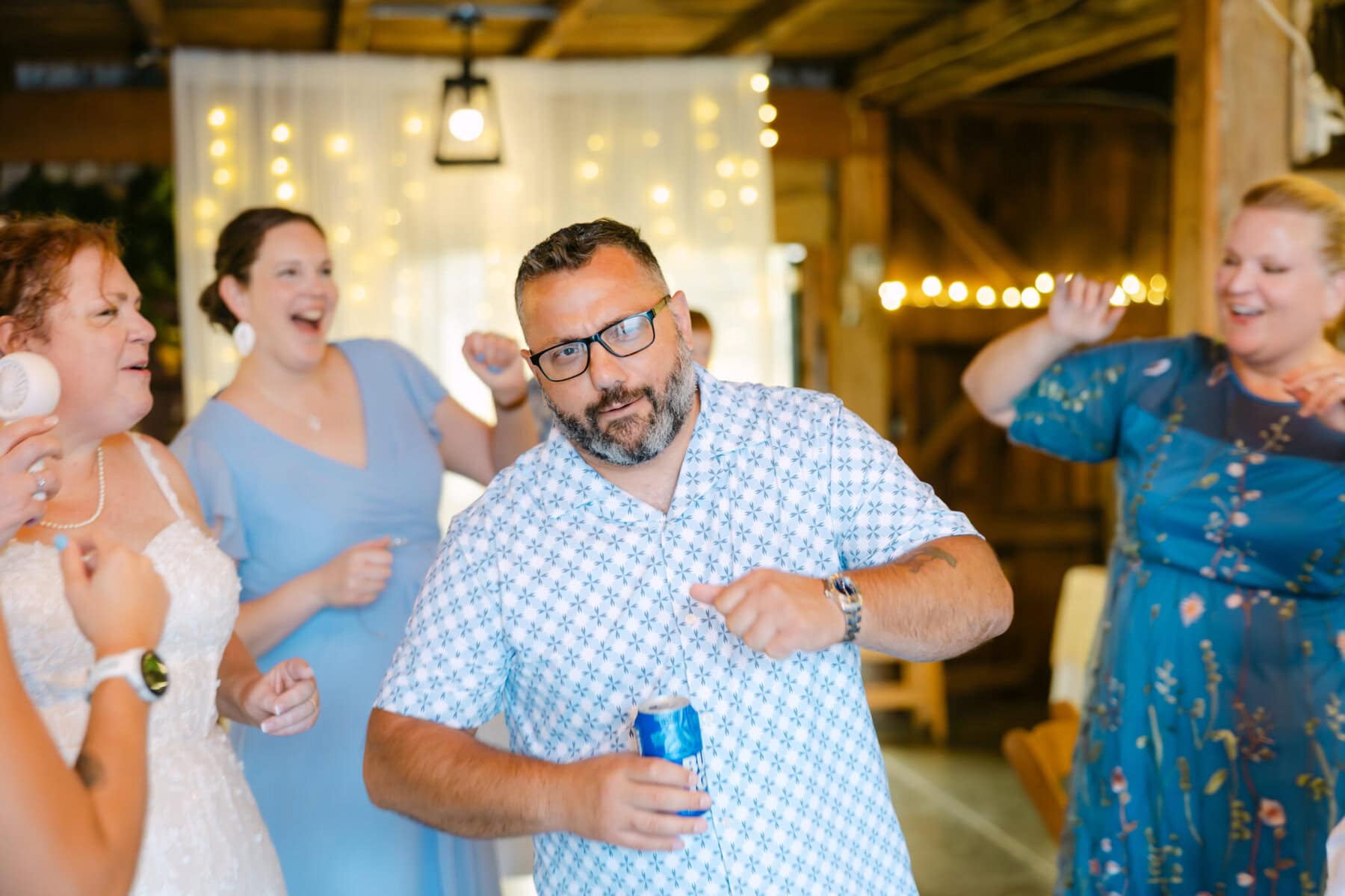 Wedding At the Holliston Historical Society 51 A man in a patterned shirt dances with a can at a wedding, surrounded by smiling women at Holliston Historical Society under string lights.