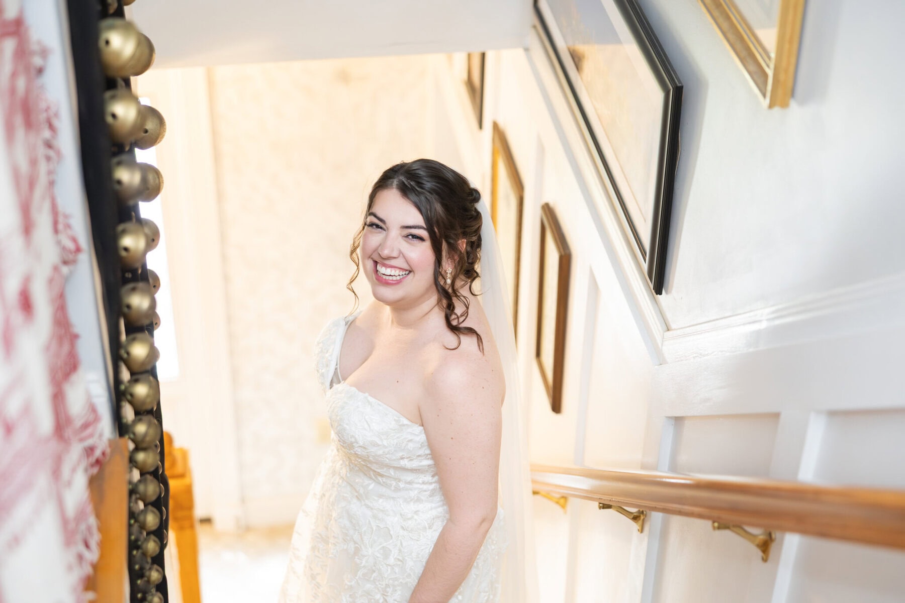 Stunning Wedding at Indian Ranch and Samuel Slater's Restaurant 9 A woman in a white wedding dress smiles on a staircase at a charming wedding venue, surrounded by framed pictures and natural light.