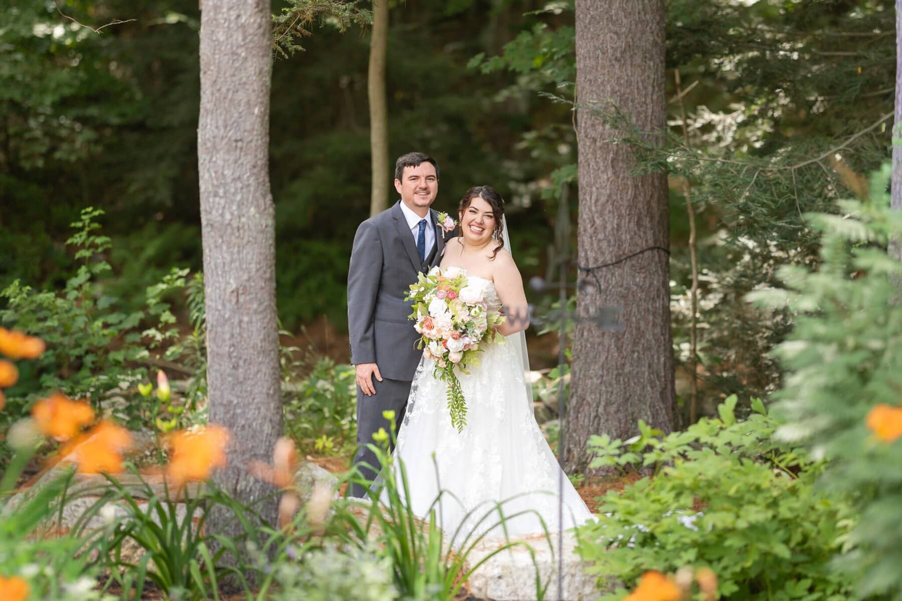 Stunning Wedding at Indian Ranch and Samuel Slater's Restaurant 22 A bride and groom stand outdoors at their wedding venue, surrounded by trees and flowers, smiling at the camera on their special day.