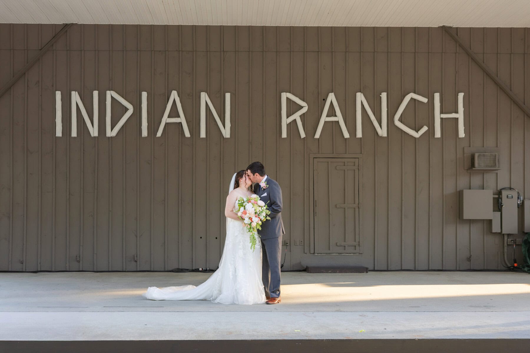 Stunning Wedding at Indian Ranch and Samuel Slater's Restaurant 112 A bride and groom share a wedding kiss in front of the large "INDIAN RANCH" letters on the building behind them.