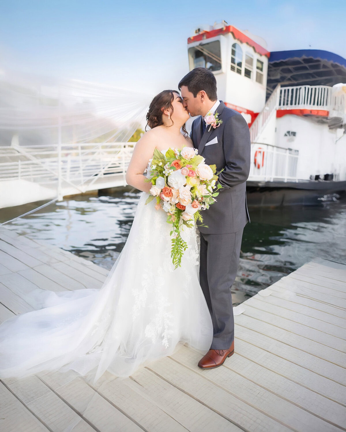 Stunning Wedding at Indian Ranch and Samuel Slater's Restaurant 63 A bride and groom stand close together on a dock at Indian Ranch, holding a bouquet, with a boat in the background.