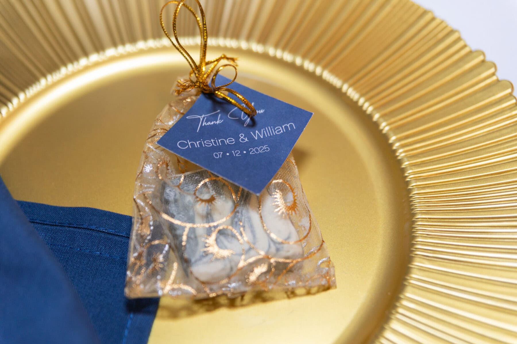 Stunning Wedding at Indian Ranch and Samuel Slater's Restaurant 74 A gold plate with a blue napkin and a wedding gift bag labeled "Christine & William 07•12•2025" at Samuel Slater's Restaurant.