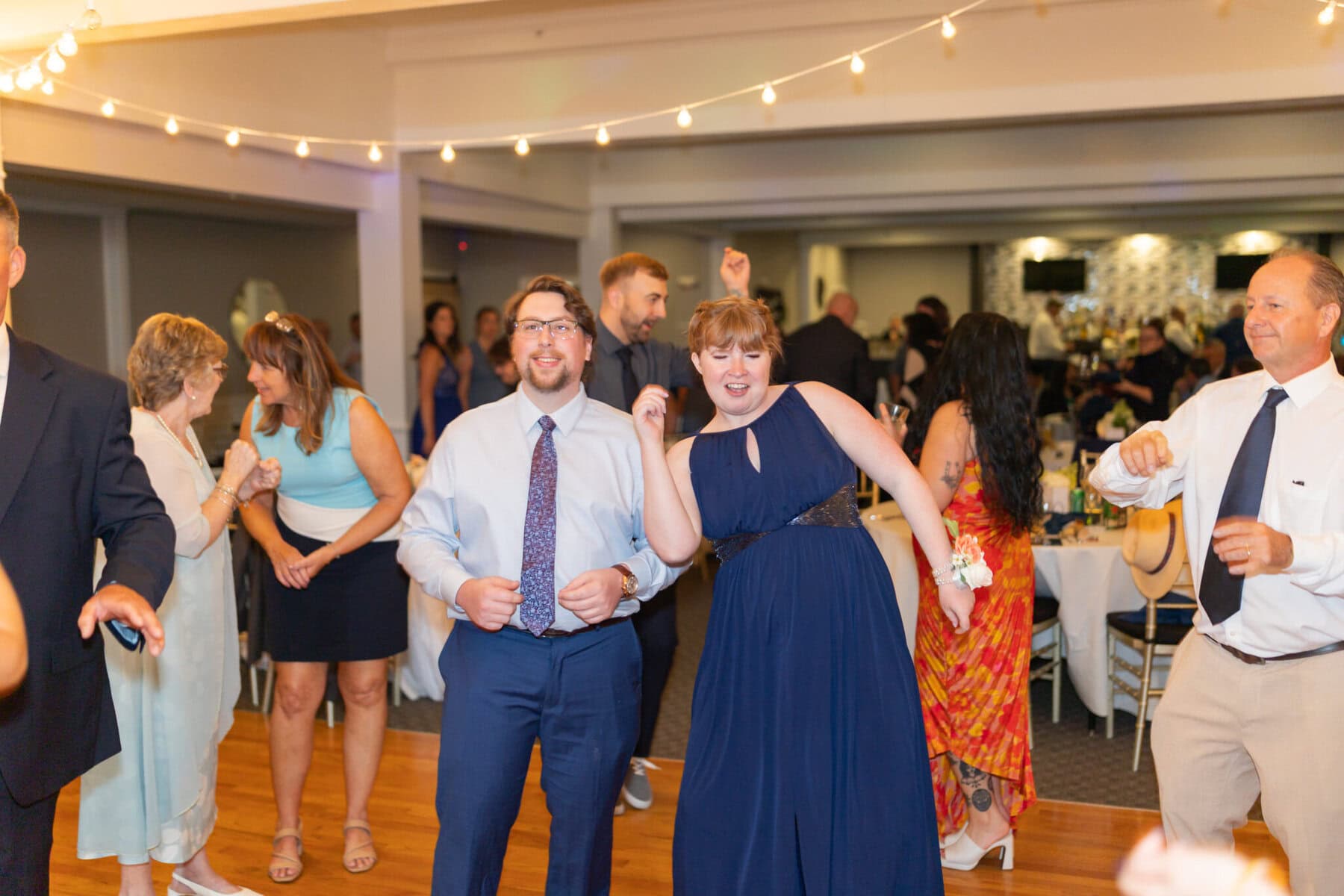 Stunning Wedding at Indian Ranch and Samuel Slater's Restaurant 89 People are dancing at a formal indoor wedding, with string lights overhead and decorated tables in the background.