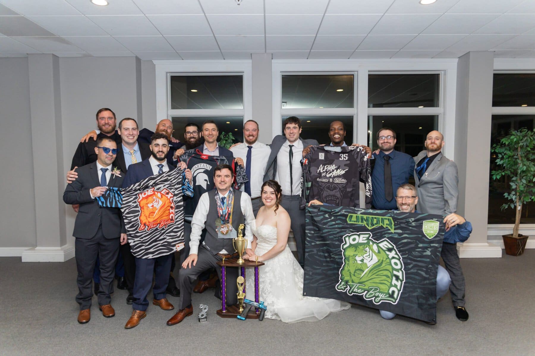Stunning Wedding at Indian Ranch and Samuel Slater's Restaurant 93 A wedding party poses indoors at Samuel Slater's Restaurant; several hold sports jerseys and a banner while the bride sits in front by trophies.