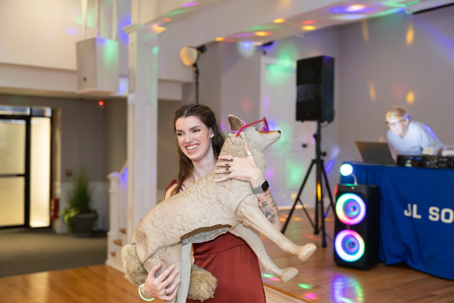 Stunning Wedding at Indian Ranch and Samuel Slater's Restaurant 100 A smiling woman in a red dress holds a large coyote statue at Samuel Slater's Restaurant, with colorful lights and a DJ booth behind her.