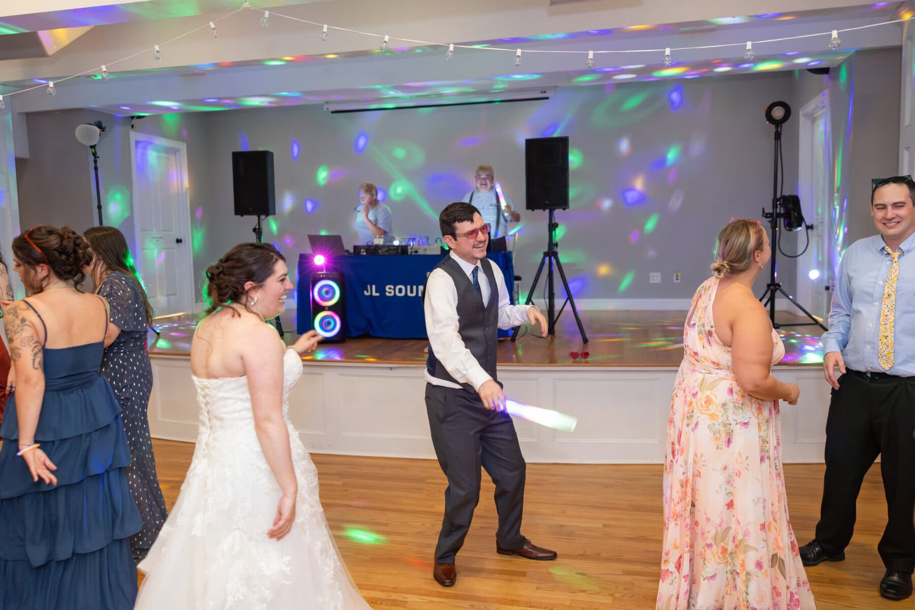 Stunning Wedding at Indian Ranch and Samuel Slater's Restaurant 103 A group of people in formal attire dance at an Indian Ranch wedding, with colorful lights and a DJ table in the decorated hall.