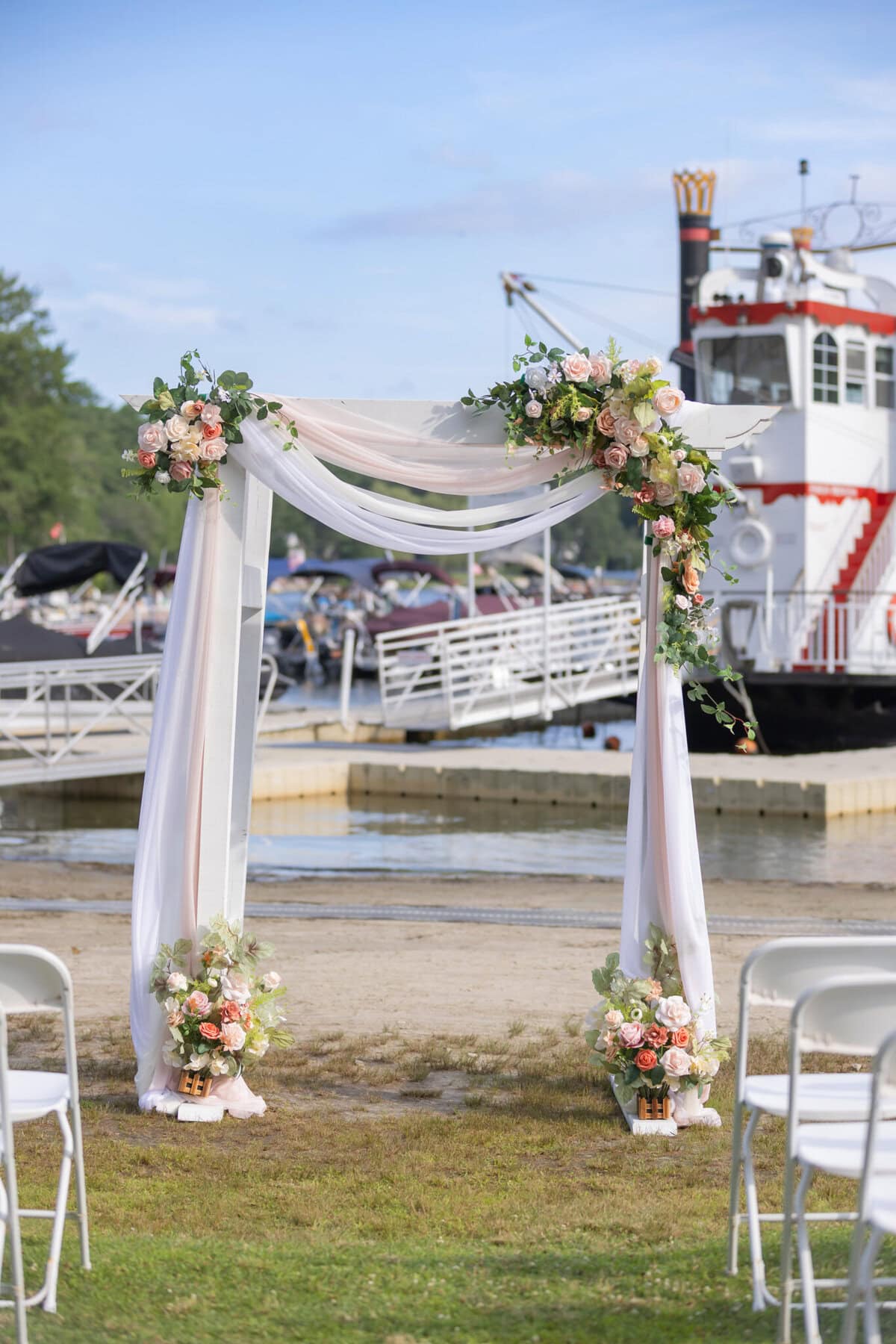 Stunning Wedding at Indian Ranch and Samuel Slater's Restaurant 27 A wedding arch with draped fabric and flowers stands by the water at Samuel Slater's Restaurant, with white chairs and a docked boat nearby.