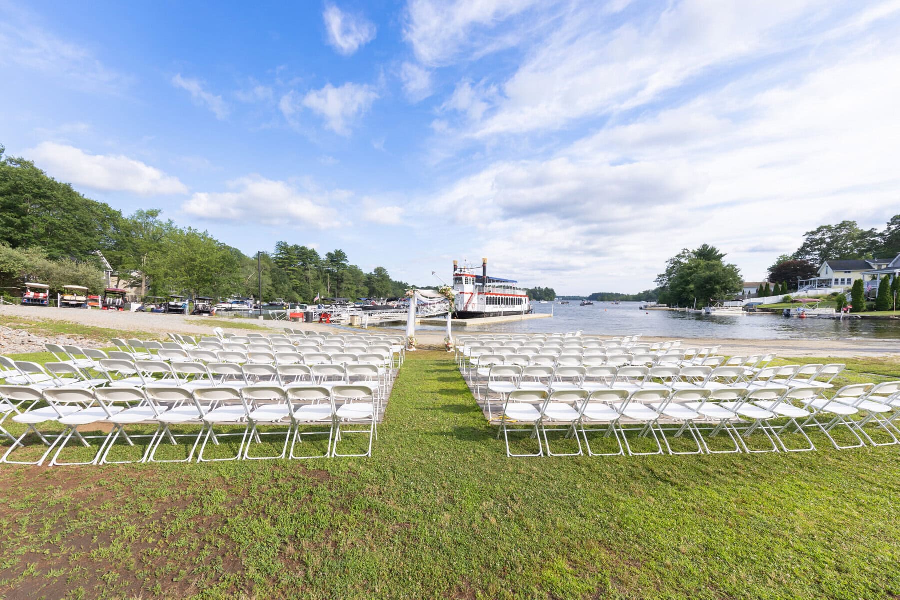 Stunning Wedding at Indian Ranch and Samuel Slater's Restaurant 28 Rows of white folding chairs set up for an Indian Ranch wedding, facing a lake near Samuel Slater's Restaurant under a partly cloudy sky.