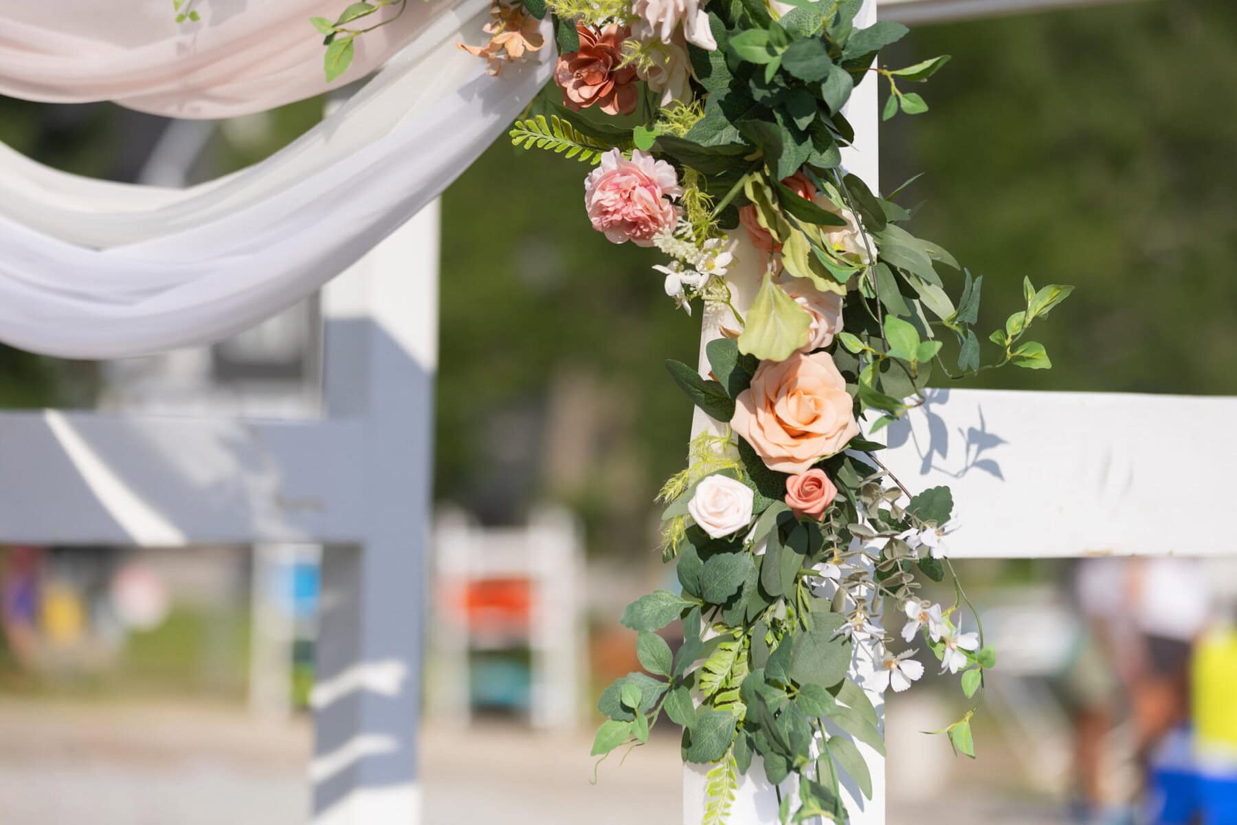 Stunning Wedding at Indian Ranch and Samuel Slater's Restaurant 29 Close-up of a white wooden wedding arch at Samuel Slater's Restaurant, adorned with pink flowers, greenery, and draped white fabric outdoors.