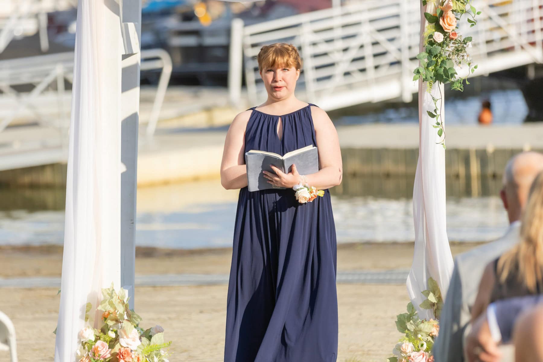 Stunning Wedding at Indian Ranch and Samuel Slater's Restaurant 32 A woman in a navy dress stands by a decorated arch at Indian Ranch, holding an open book, with water and boats in the background.