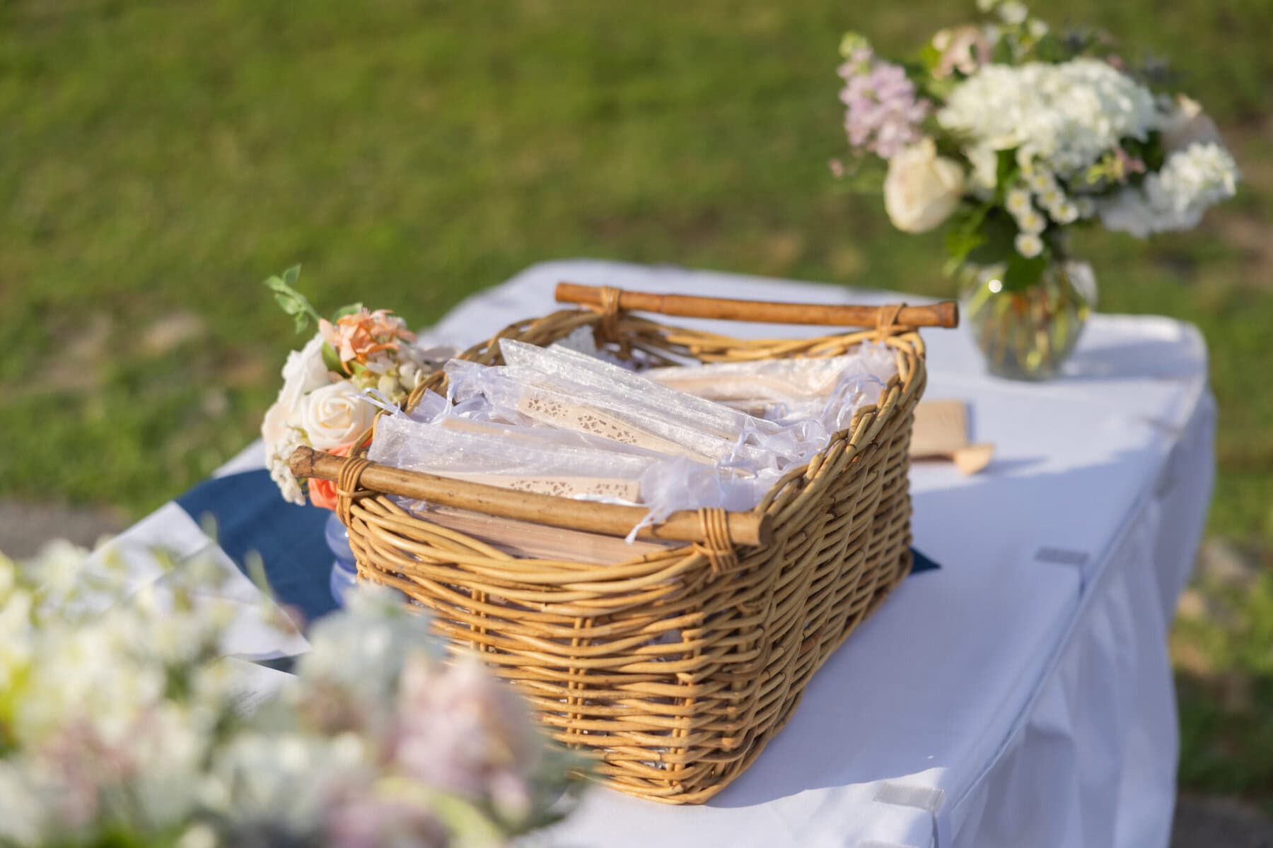 Stunning Wedding at Indian Ranch and Samuel Slater's Restaurant 30 A wicker basket with lace-wrapped items sits on a white table outdoors at a wedding, surrounded by floral arrangements.