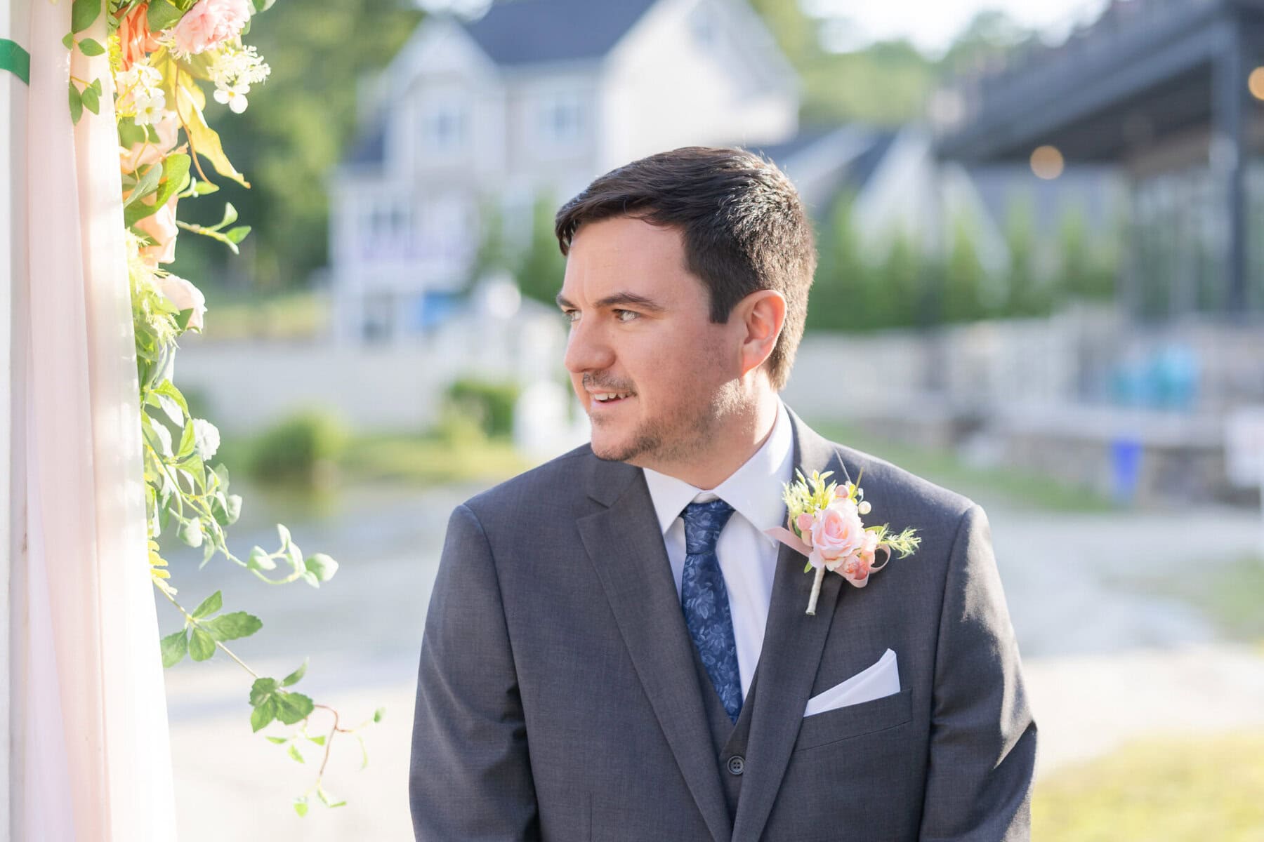 Stunning Wedding at Indian Ranch and Samuel Slater's Restaurant 33 A man in a suit with a pink boutonniere stands outdoors at Samuel Slater's Restaurant wedding, blurred buildings and greenery behind him.