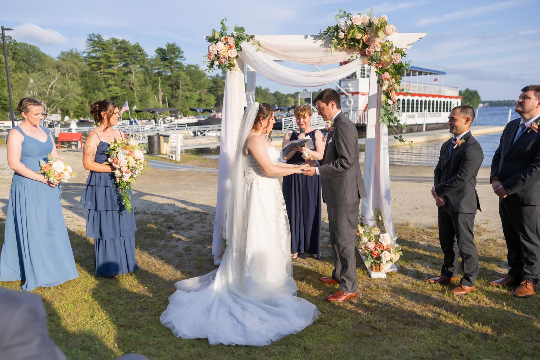 Stunning Wedding at Indian Ranch and Samuel Slater's Restaurant 38 A bride and groom exchange vows under a floral arch by the water at Samuel Slater's Restaurant, surrounded by their wedding party.