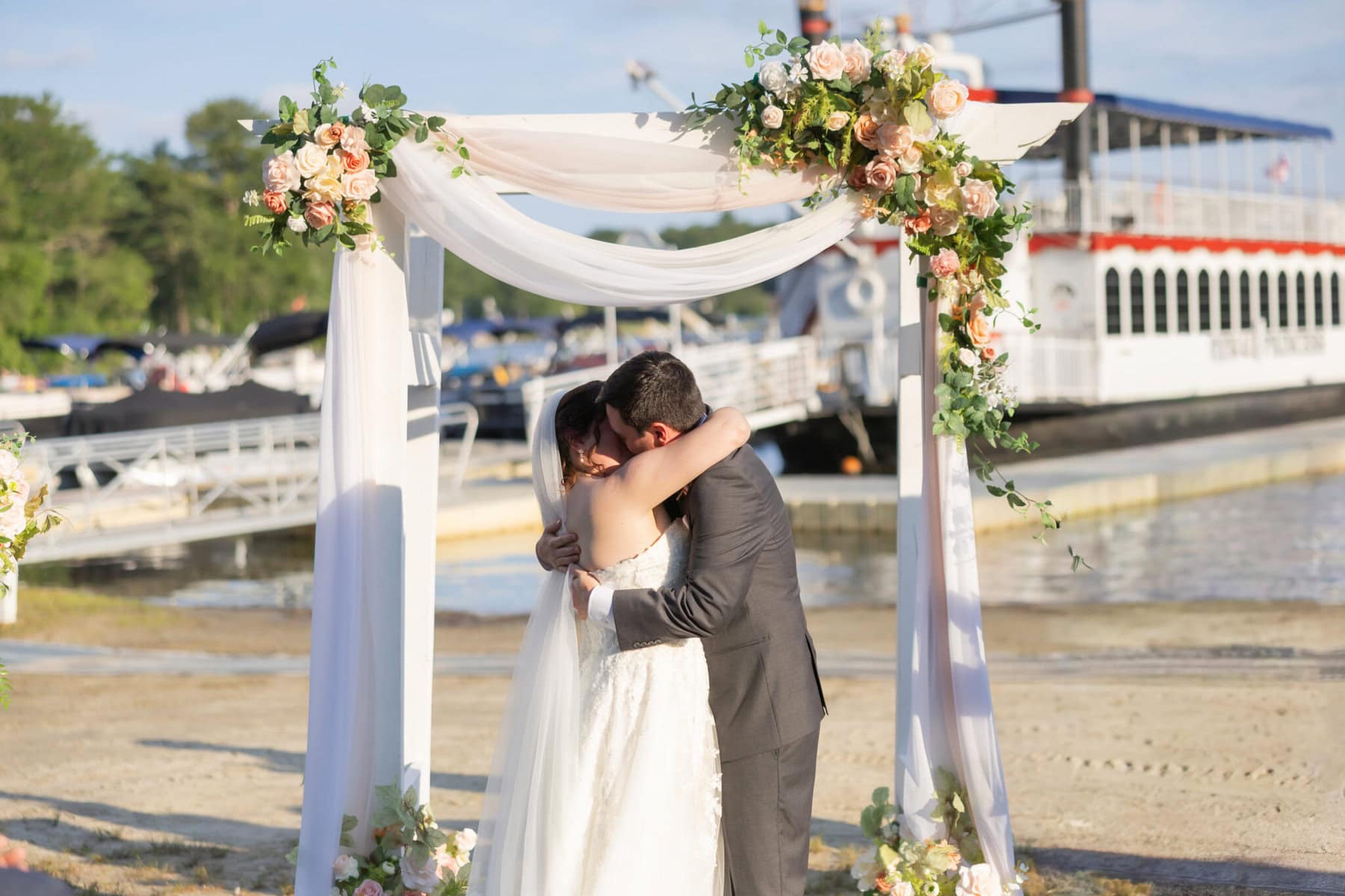 Stunning Wedding at Indian Ranch and Samuel Slater's Restaurant 44 A bride and groom embrace under a flower arch by the water at Indian Ranch, with a large boat docked in the background.