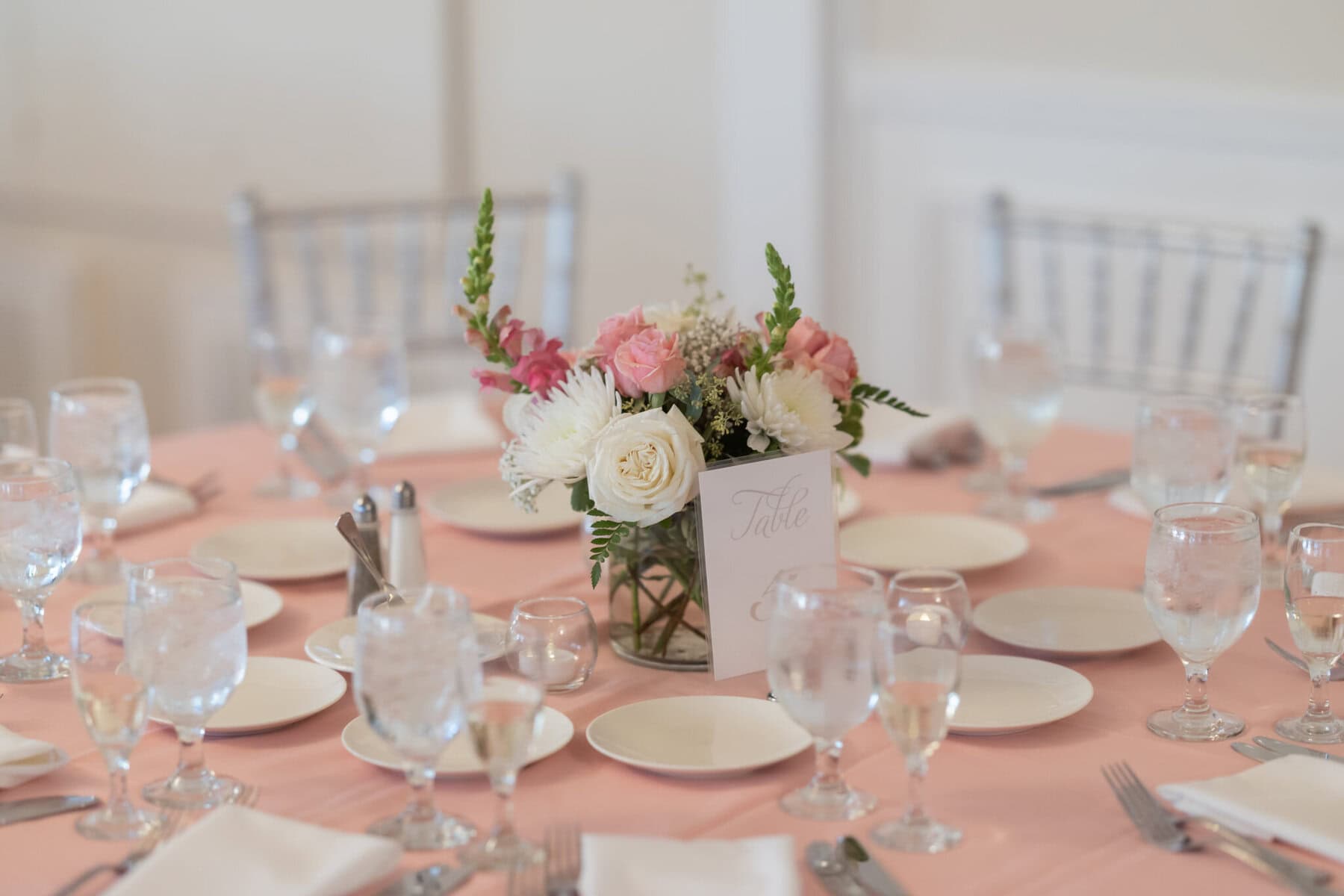 Beautiful Lake Pearl Wedding in Wrentham, MA 25 A round table with a pink tablecloth, set for a Lake Pearl Wedding in Wrentham MA, features plates, glasses, and a floral centerpiece.
