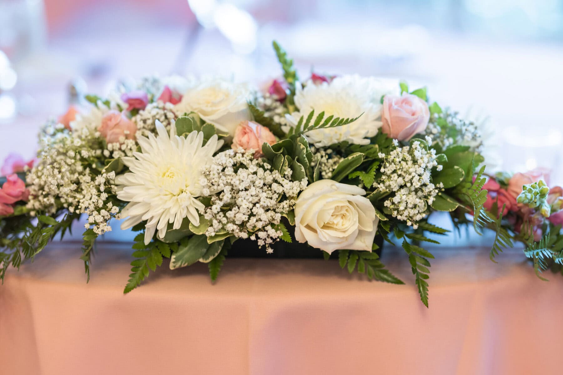 Beautiful Lake Pearl Wedding in Wrentham, MA 24 Floral arrangement with white chrysanthemums, roses, baby’s breath, and green ferns adorns a pink tablecloth at a Lake Pearl Wedding in Wrentham MA.