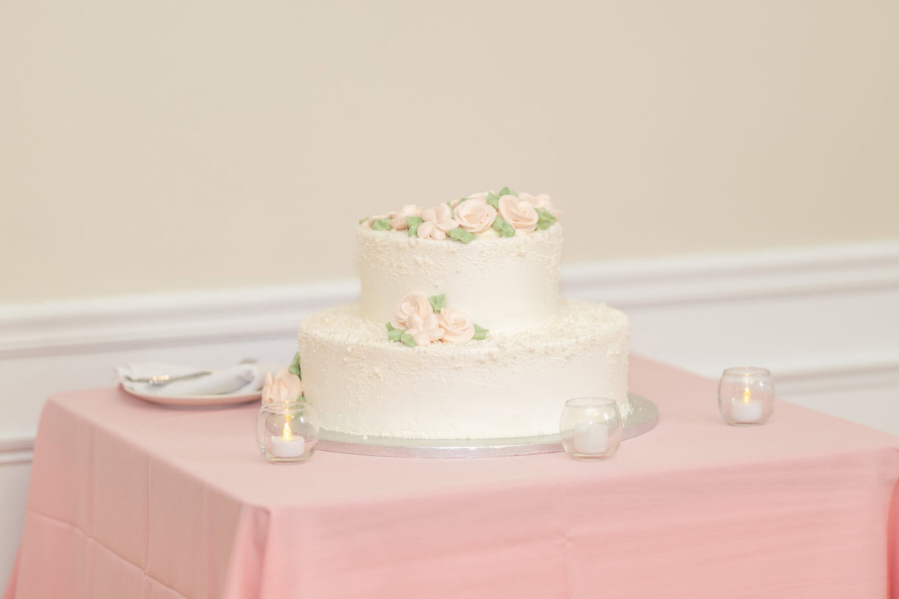 Beautiful Lake Pearl Wedding in Wrentham, MA 20 A two-tier white cake with pink roses graces a Lake Pearl Wedding in Wrentham MA, set on a pink tablecloth amid candles and plates.