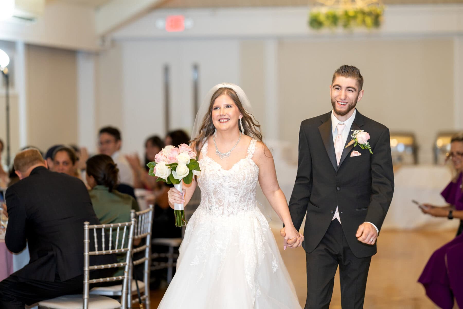 Beautiful Lake Pearl Wedding in Wrentham, MA 32 A bride and groom walk hand in hand, smiling at their Lake Pearl Wedding in Wrentham, MA, with guests seated nearby.