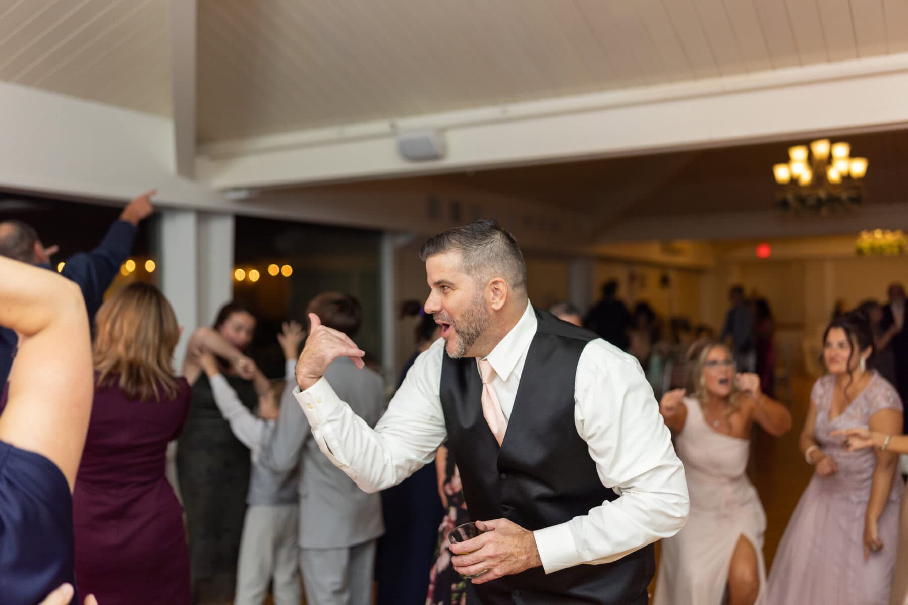 Beautiful Lake Pearl Wedding in Wrentham, MA 42 A man in a vest and tie dances enthusiastically at a Lake Pearl Wedding in Wrentham MA, with other guests dancing in the background.