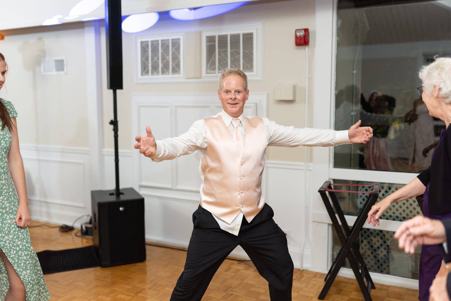 Beautiful Lake Pearl Wedding in Wrentham, MA 45 A man in a light pink vest and tie stands with arms wide open on the Lake Pearl Wedding dance floor in Wrentham, MA, surrounded by people.