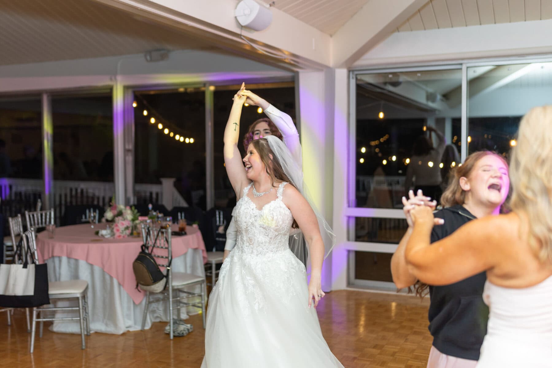 Beautiful Lake Pearl Wedding in Wrentham, MA 46 A bride in a white dress dances with her partner indoors at a Lake Pearl Wedding in Wrentham, MA; guests sway beneath string lights.