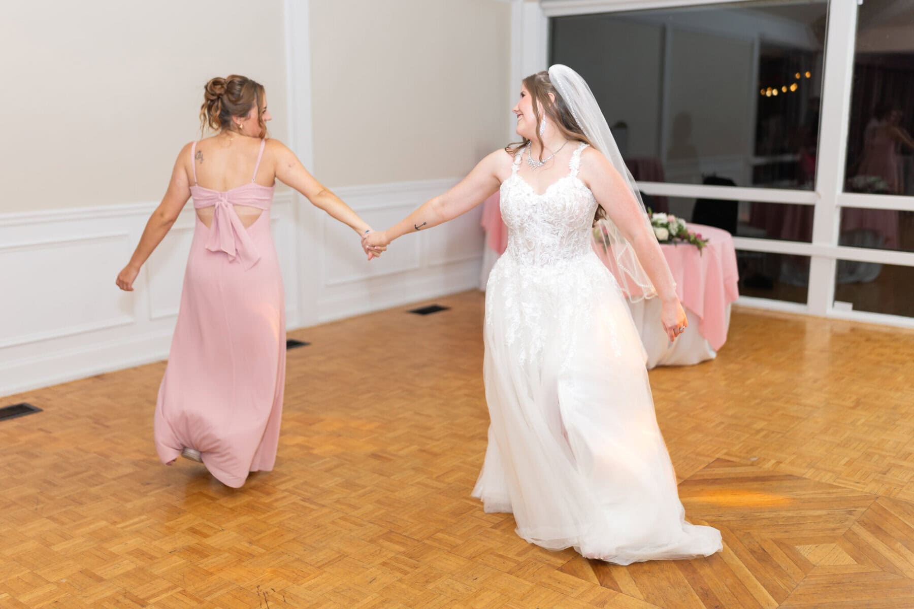 Beautiful Lake Pearl Wedding in Wrentham, MA 47 A bride in a white dress and veil dances with her bridesmaid on a wooden floor at their Lake Pearl Wedding in Wrentham, MA.