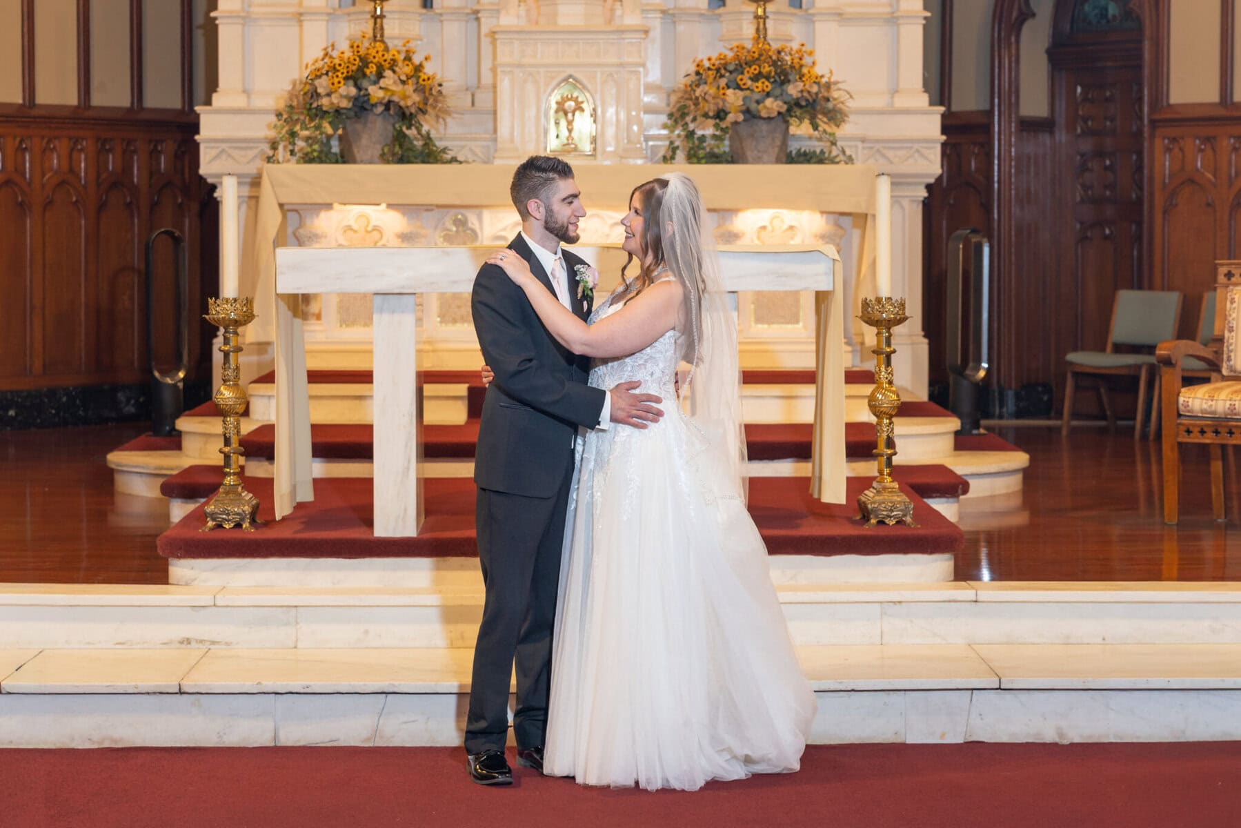 Wedding At St. Mary's Church in North Attleborough 2 A bride and groom stand smiling at each other inside St. Mary's Church, in front of a flower-decorated altar in North Attleborough.
