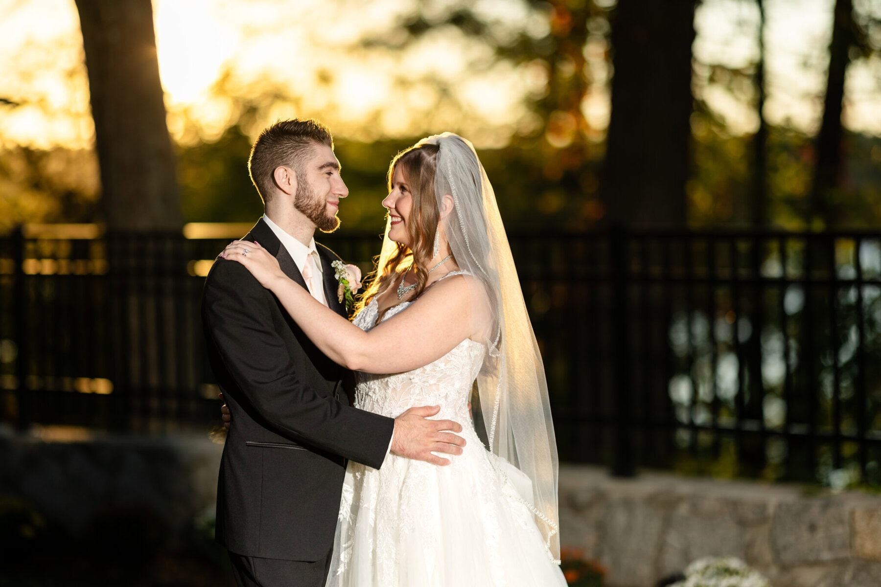 Beautiful Lake Pearl Wedding in Wrentham, MA 4 A bride and groom stand outdoors at sunset during their Lake Pearl Wedding, smiling with trees and a black fence in the Wrentham MA background.