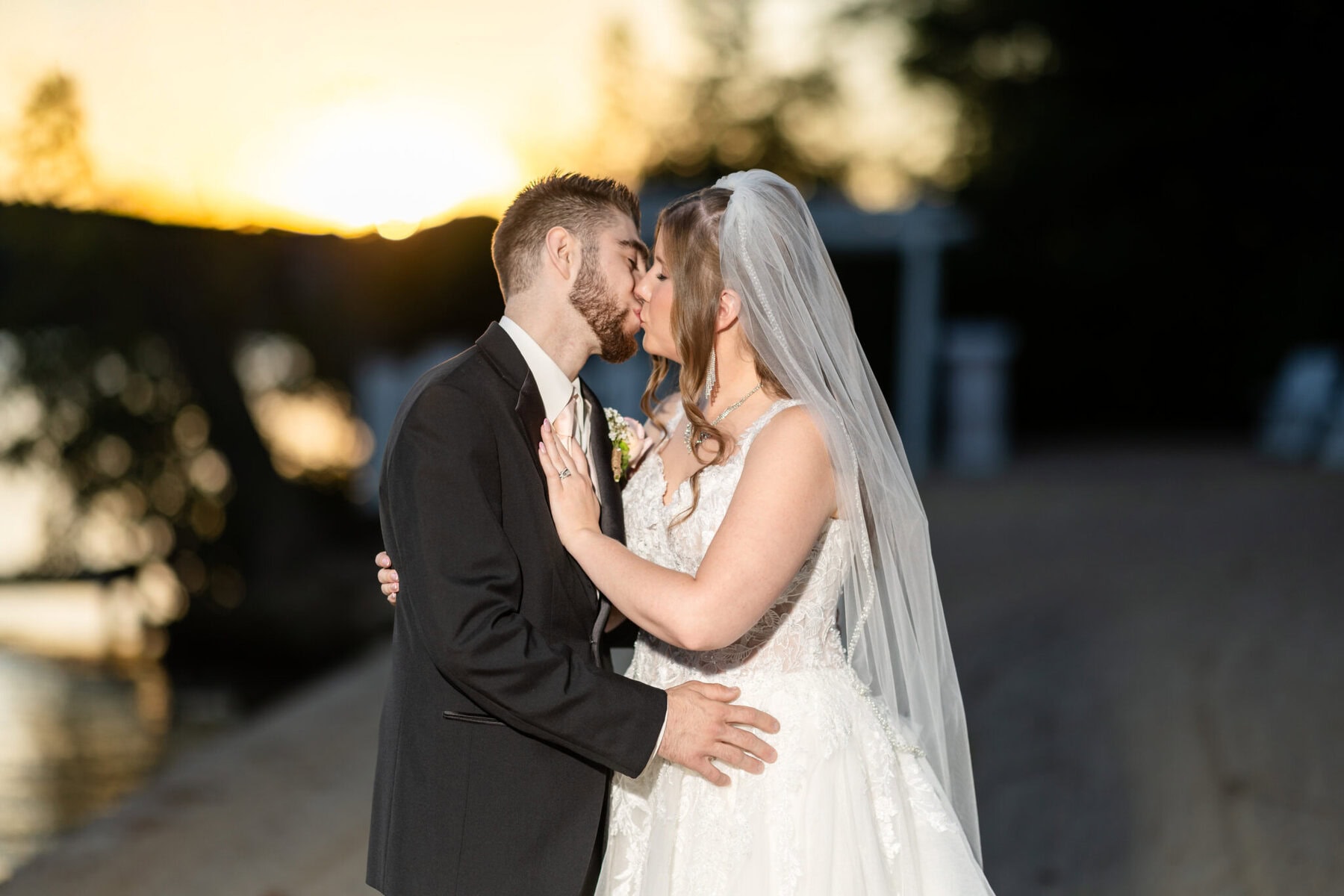 Beautiful Lake Pearl Wedding in Wrentham, MA 8 A bride and groom kiss at sunset by Lake Pearl, Wrentham MA—she in a white dress and veil, he in a classic black suit.