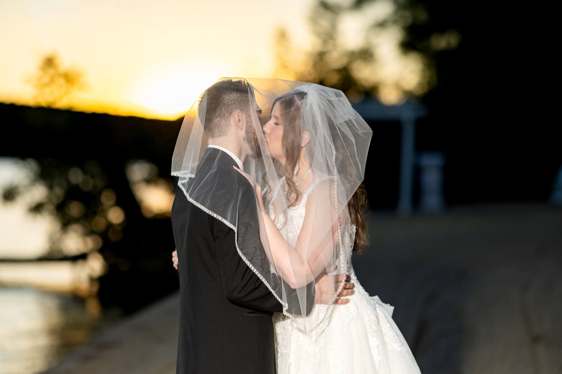 Beautiful Lake Pearl Wedding in Wrentham, MA 9 A bride and groom kiss outdoors at sunset at their Lake Pearl Wedding in Wrentham, MA, the bride’s veil softly covering their faces.