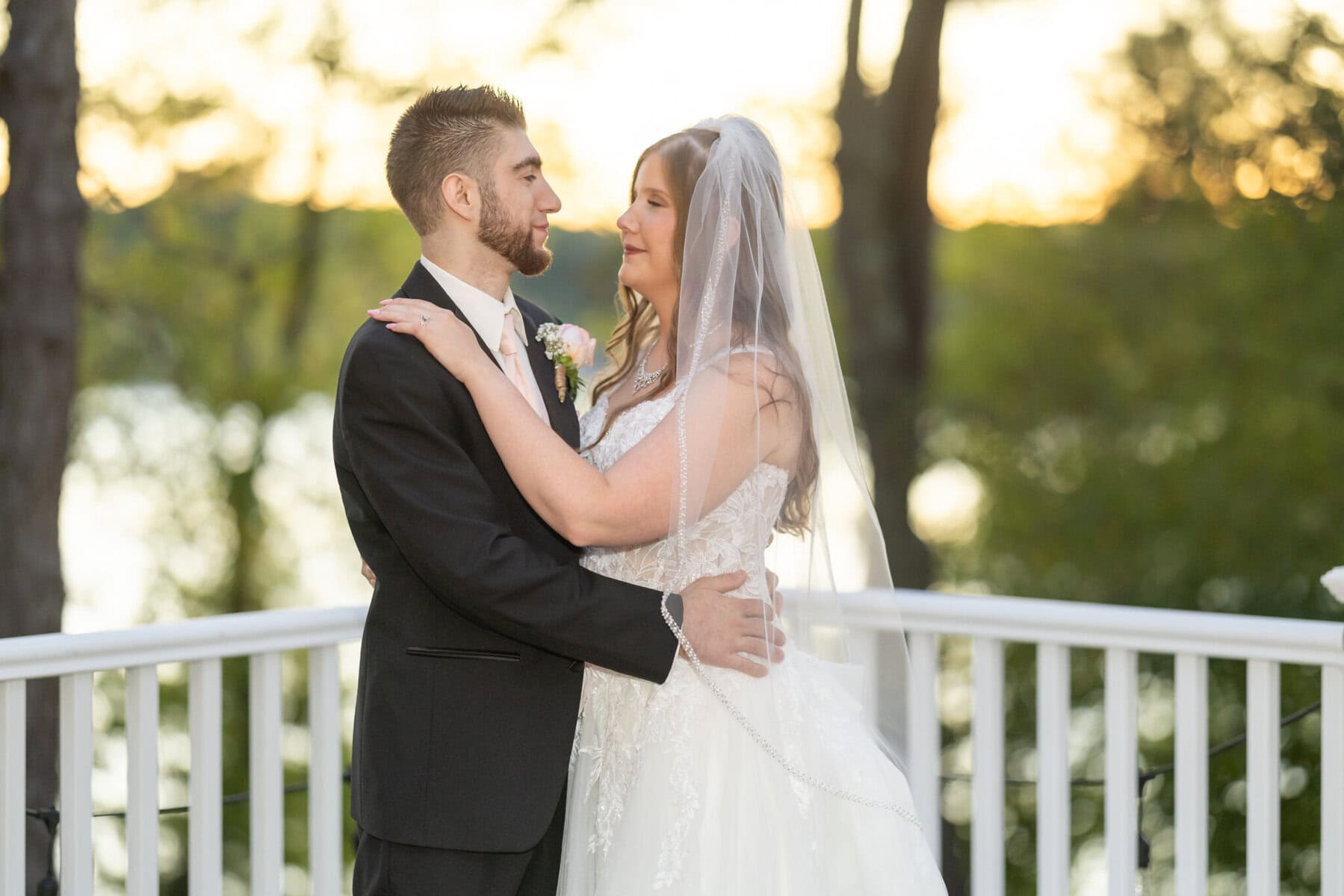 Beautiful Lake Pearl Wedding in Wrentham, MA 12 A bride and groom embrace on a white outdoor balcony with greenery in the background at their Lake Pearl Wedding in Wrentham, MA.