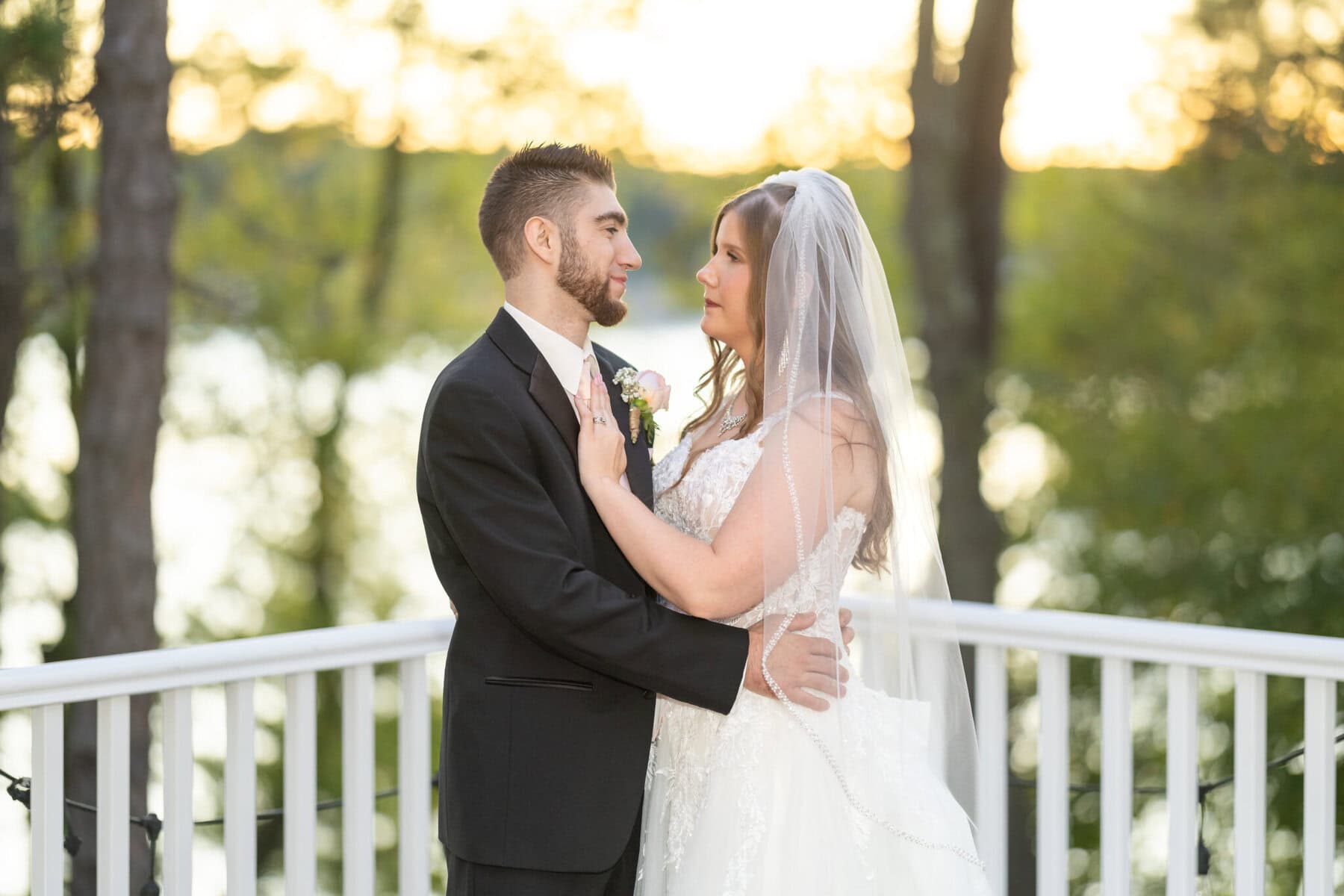 Beautiful Lake Pearl Wedding in Wrentham, MA 13 A bride and groom stand on a white railing outdoors at their Lake Pearl Wedding, with trees and water in the background.