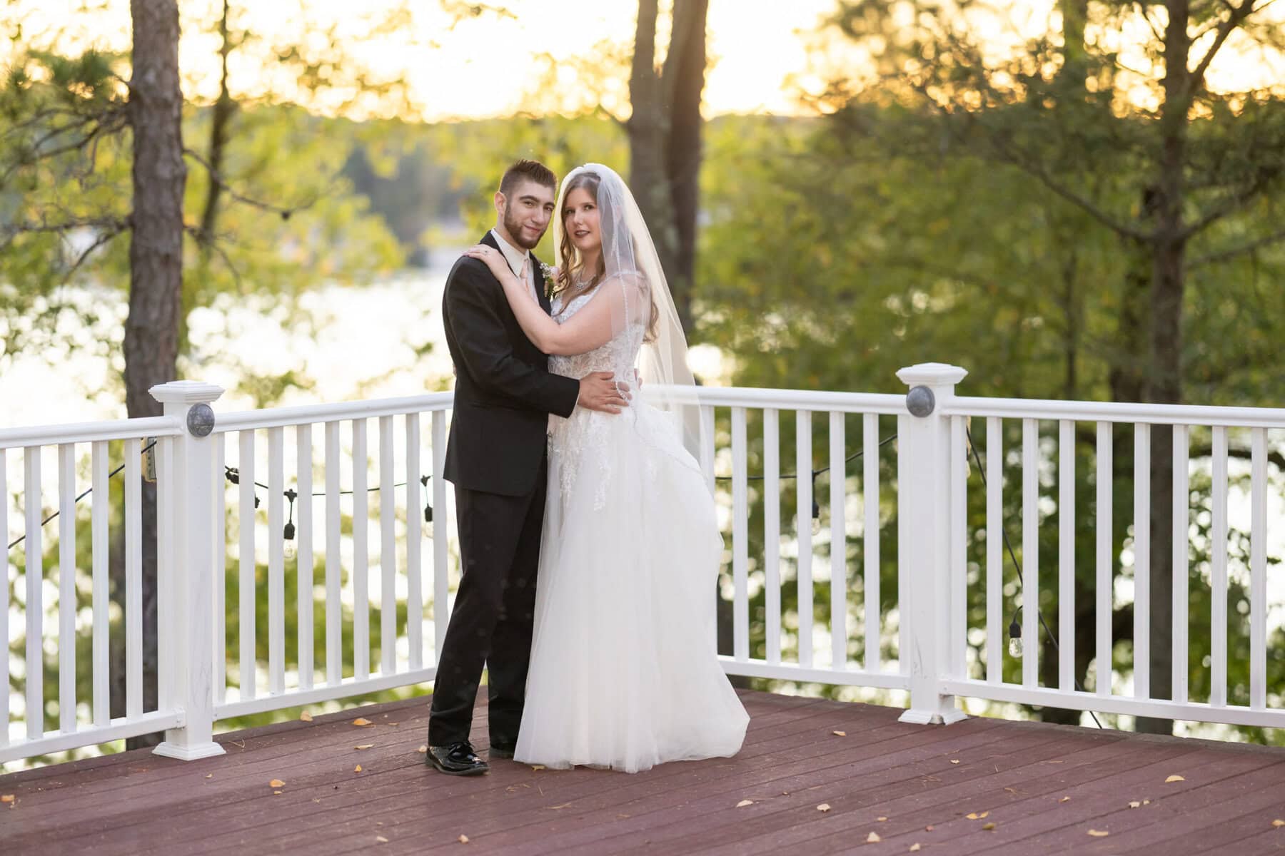 Beautiful Lake Pearl Wedding in Wrentham, MA 14 A bride and groom in formal attire stand on a wooden deck at their Lake Pearl Wedding, surrounded by trees and sunlight in Wrentham MA.