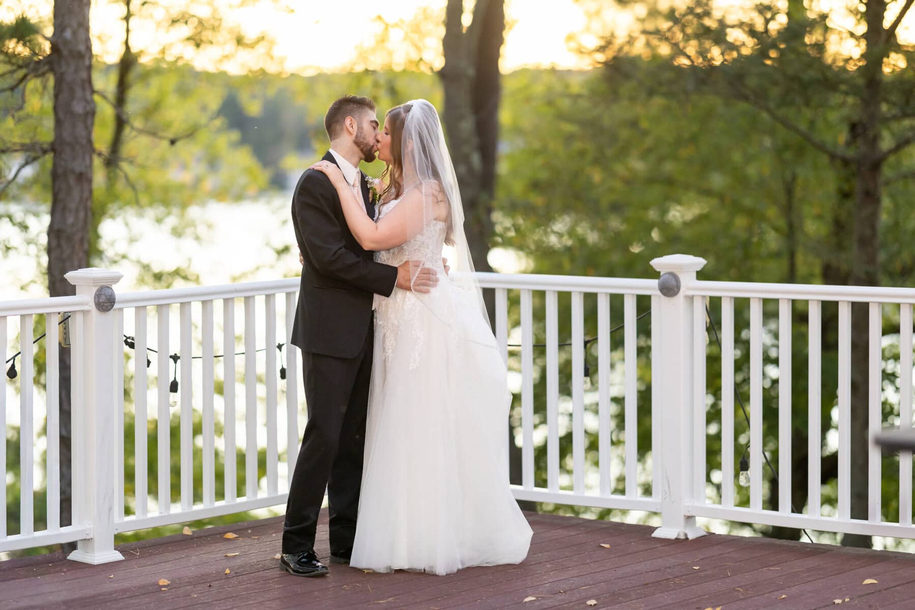 Beautiful Lake Pearl Wedding in Wrentham, MA 11 A bride and groom embrace and kiss on a wooden deck outdoors at their Lake Pearl Wedding in Wrentham, MA, with trees and water behind them.