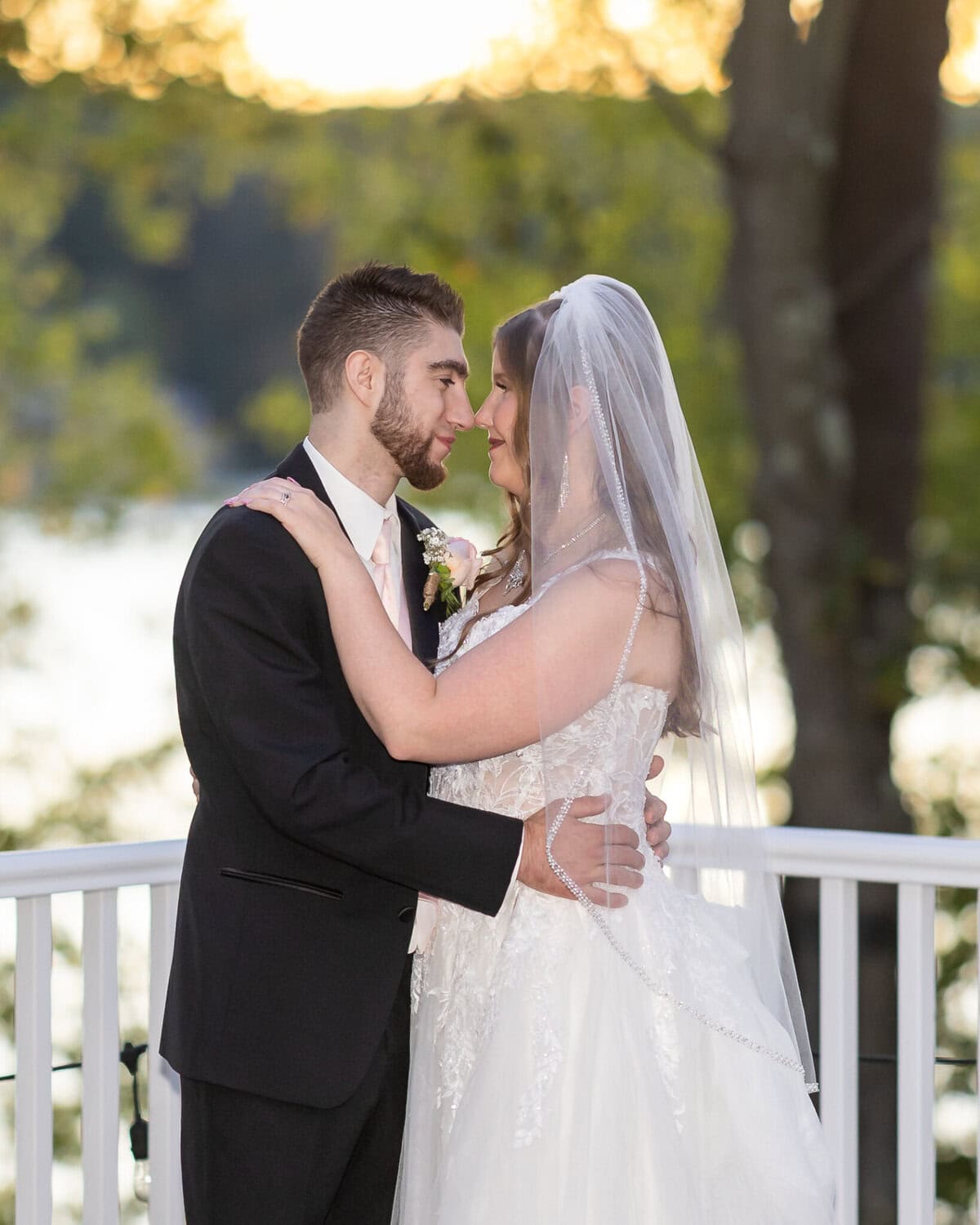 Beautiful Lake Pearl Wedding in Wrentham, MA 18 A bride and groom embrace by a white railing at their Lake Pearl Wedding in Wrentham, MA, smiling with trees and water behind them.