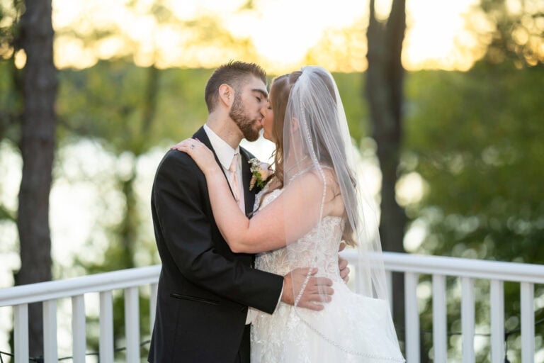 A bride and groom in wedding attire kiss outdoors by a white railing at their Lake Pearl Wedding in Wrentham, MA, sunlight streaming through trees.