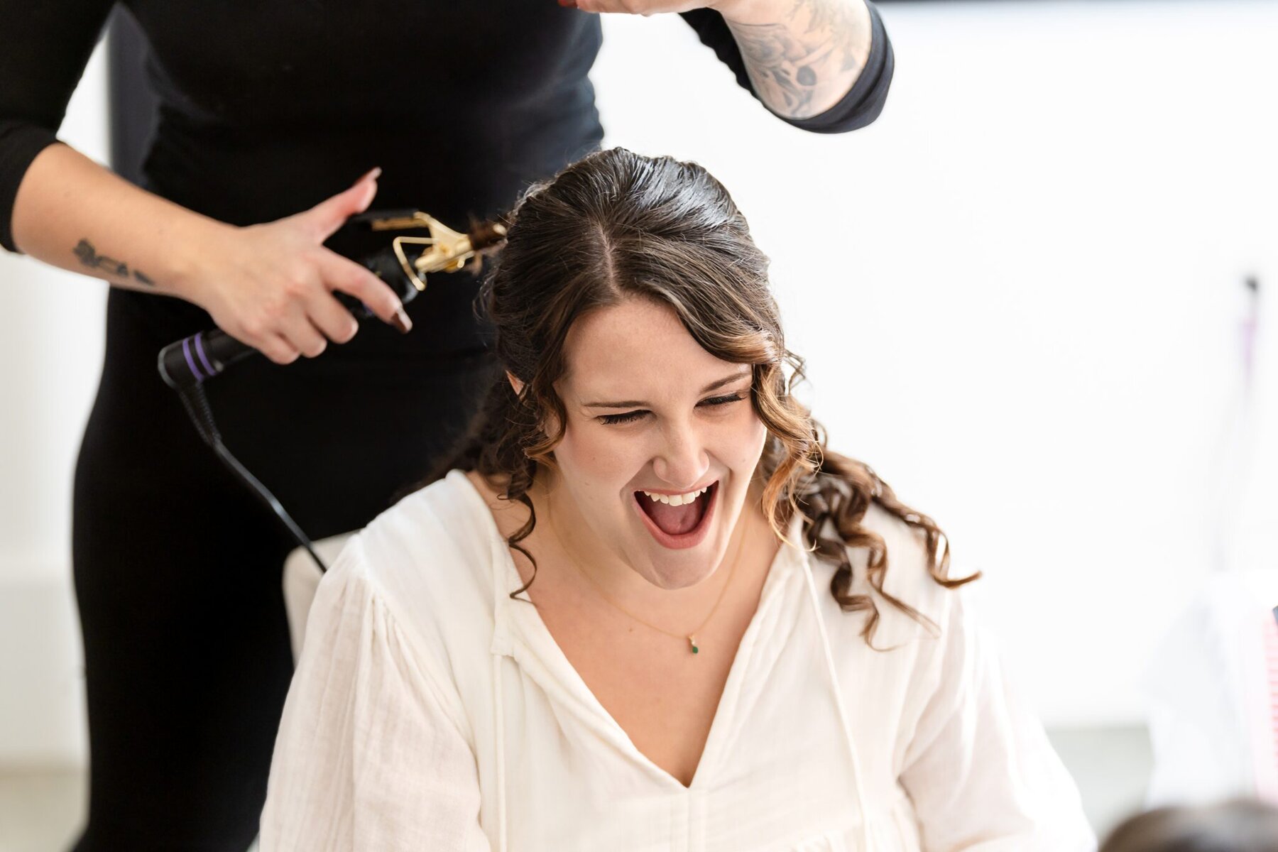 Wedding at the Newport Naval Station Officers' Club 2 A woman with wavy hair smiles widely as another prepares her for a wedding at the Officers' Club with a curling iron.