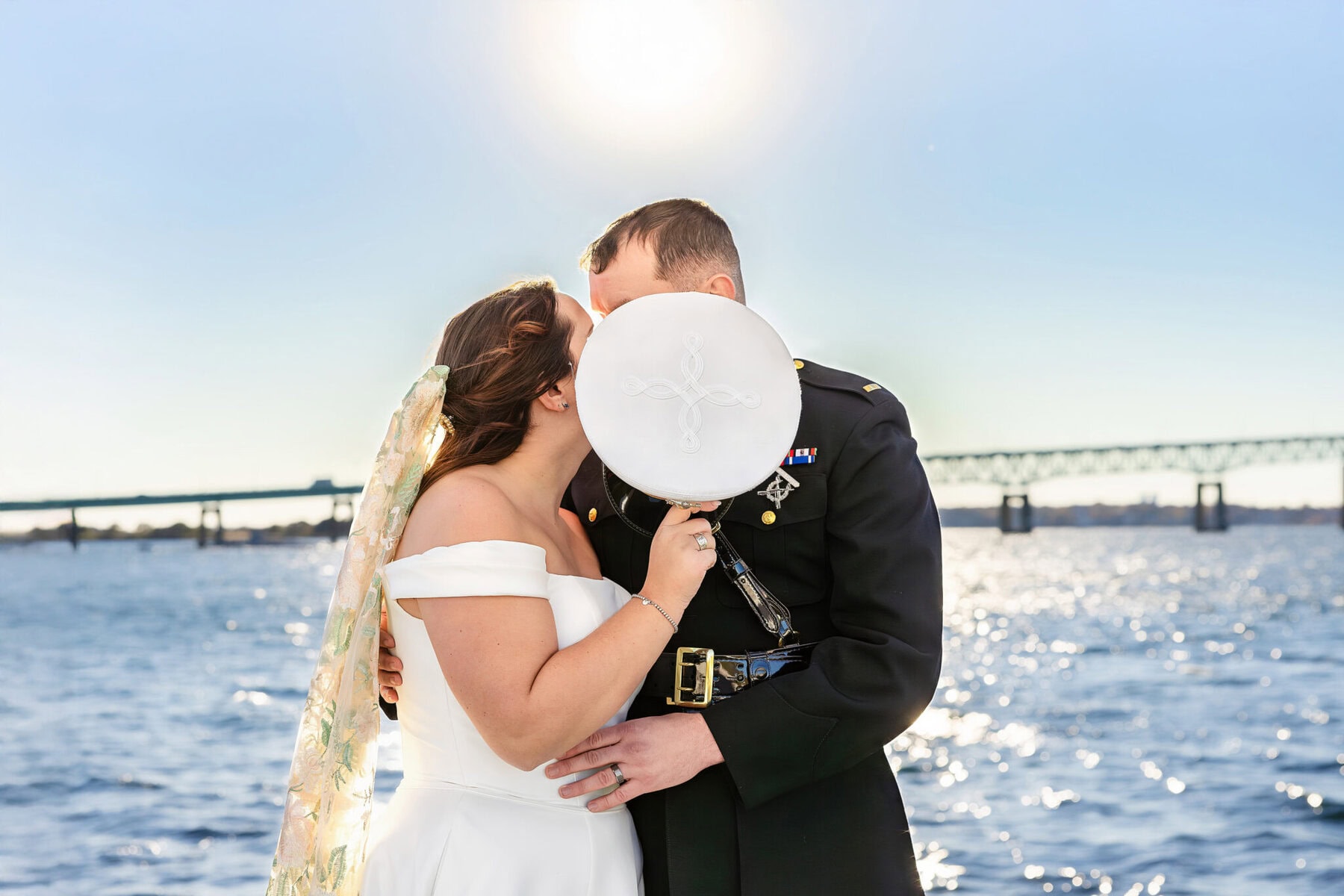 Wedding at the Newport Naval Station Officers' Club 48 A bride and groom in formal attire share a secret kiss behind a white military hat at the Officers' Club, Newport Naval Station.