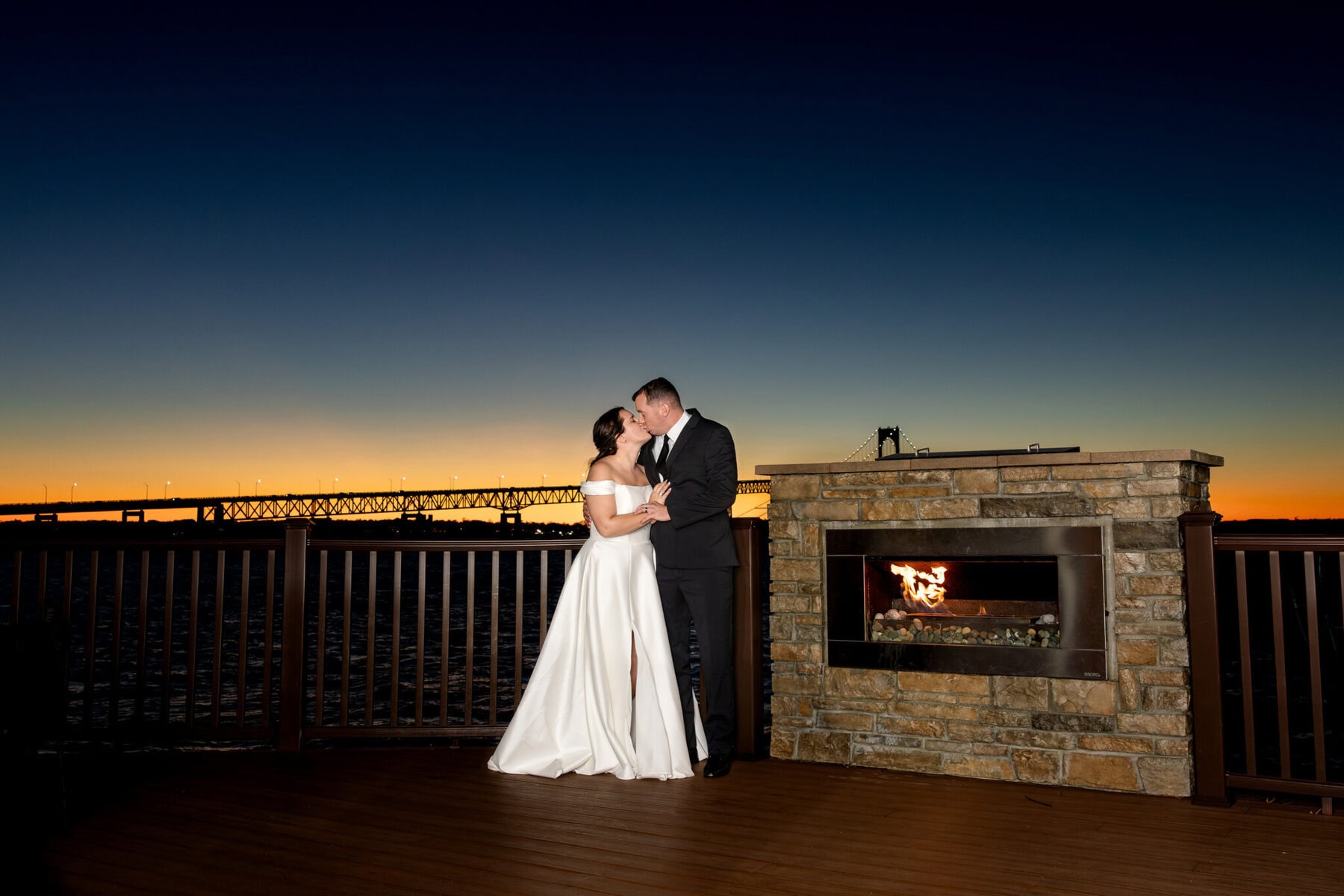 Wedding at the Newport Naval Station Officers' Club 112 A bride and groom stand by an outdoor fireplace at sunset on the Officers' Club deck, with Newport Naval Station's bridge in the background.